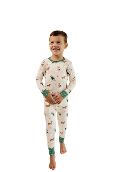 A young boy smiling and walking forward while wearing a cream-colored long-sleeve pajama set with green cuffs and hem, decorated with a dog print, against a plain white studio background.