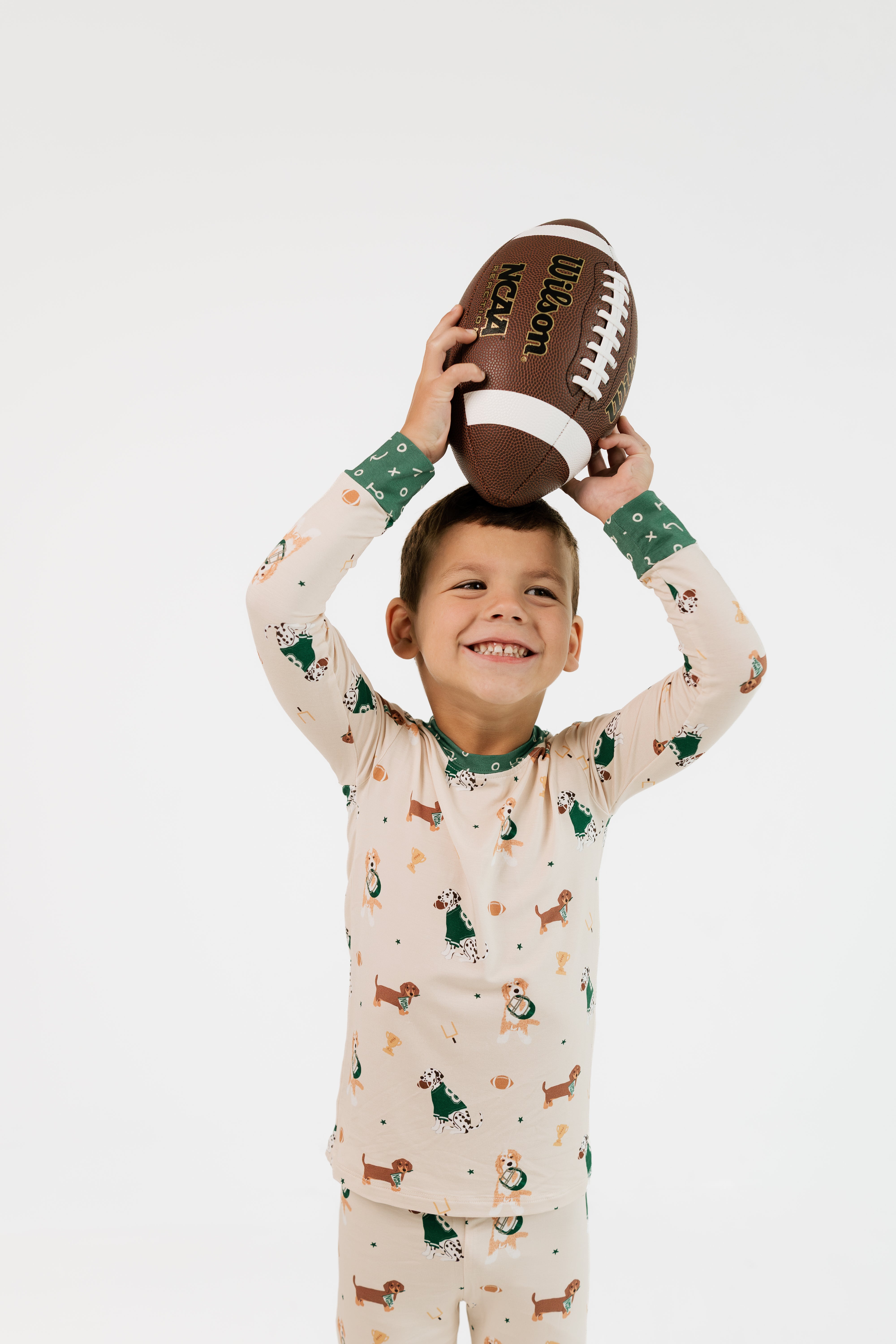 A young boy smiling and holding a brown football above his head, wearing a long-sleeve pajama set with a dachshund print in green, brown, and orange on a cream background. The photo is shot against a seamless white studio background.