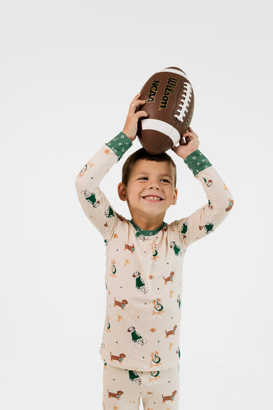A young boy smiling and holding a brown football above his head, wearing a long-sleeve pajama set with a dachshund print in green, brown, and orange on a cream background. The photo is shot against a seamless white studio background.