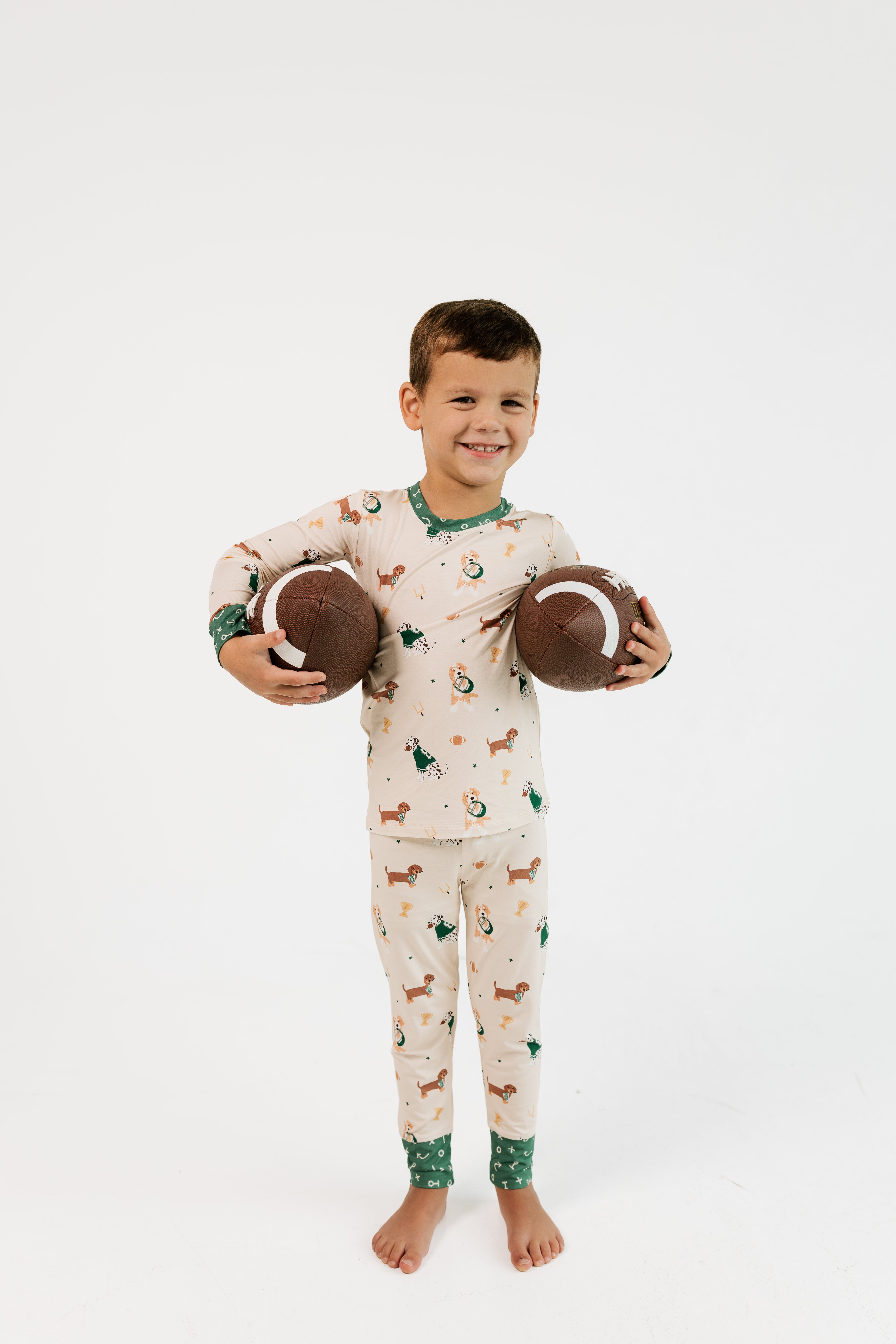 A young boy stands barefoot against a plain white studio background, holding two brown footballs. He is wearing a long-sleeve pajama set with a woodland dog print on a cream base and green trim at the collar, cuffs, and ankles.