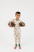 A young boy stands barefoot against a plain white studio background, holding two brown footballs. He is wearing a long-sleeve pajama set with a woodland dog print on a cream base and green trim at the collar, cuffs, and ankles.