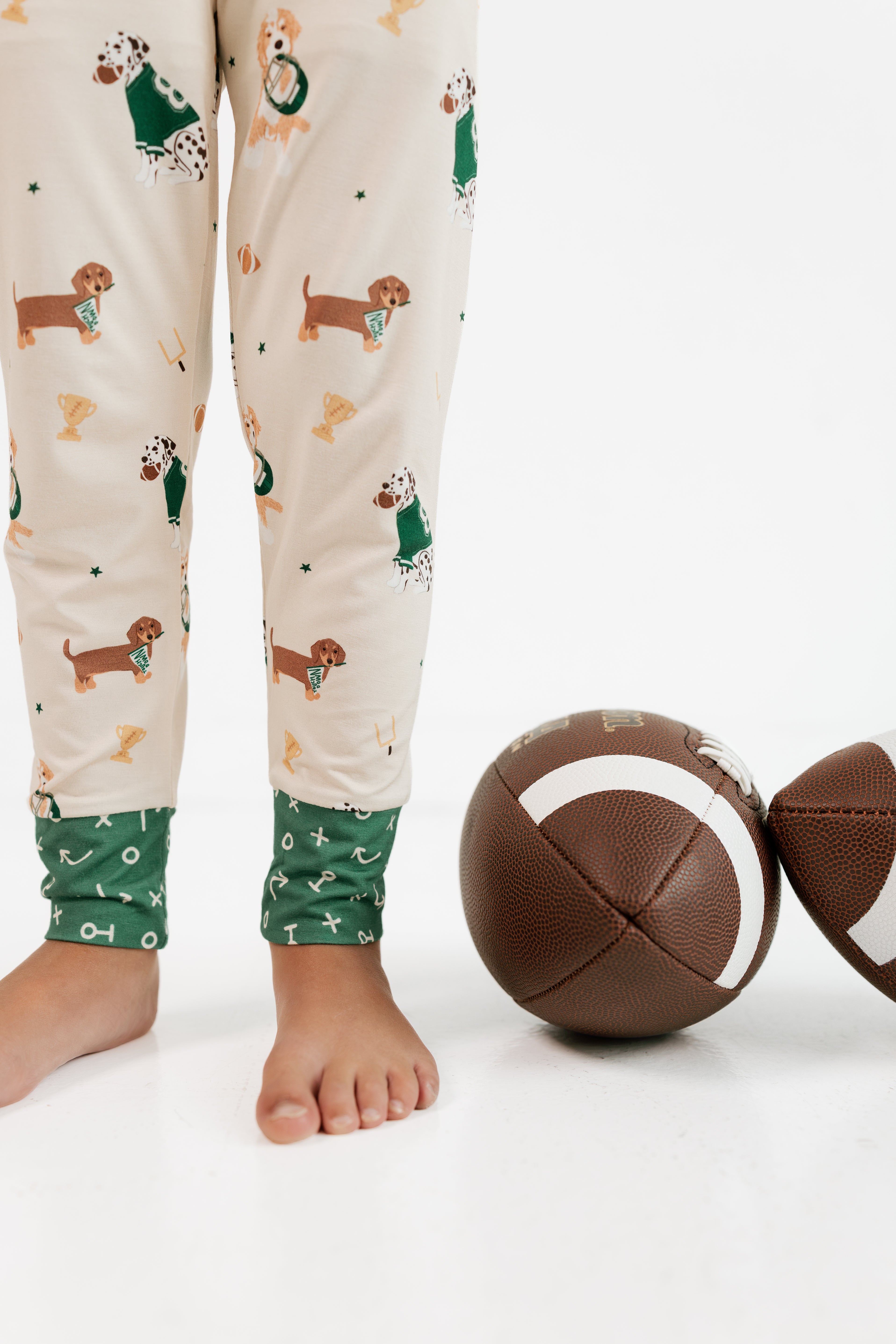 A person wearing cream pajama pants with a dachshund and green sweater print, standing barefoot next to two footballs on a white studio floor.