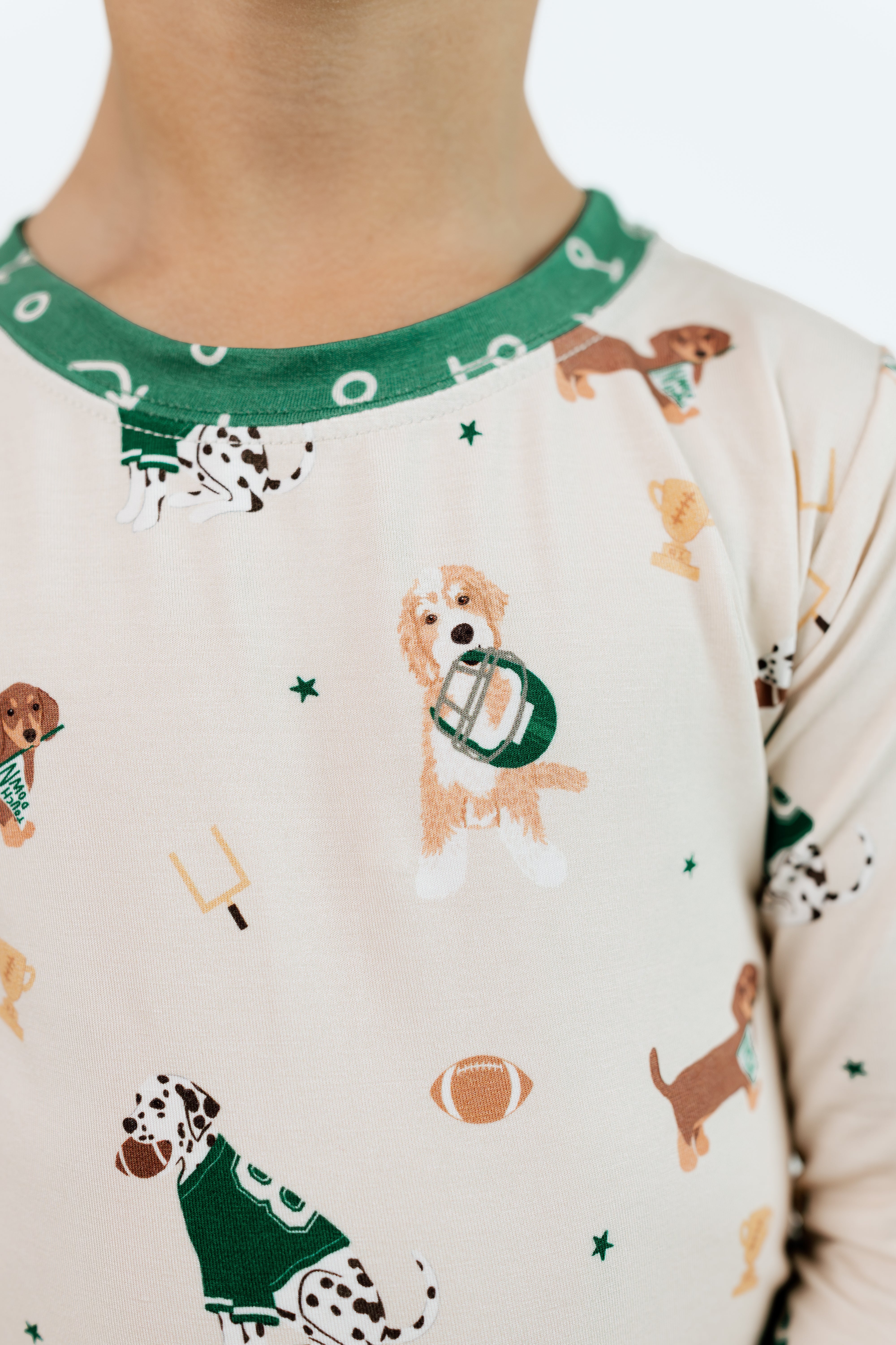 A close-up of a child wearing a long-sleeve pajama top with a cream background and a green collar. The pajama top features an all-over print of dalmatian and golden retriever dogs in football uniforms, along with football-themed icons like a helmet, football, and tackles.