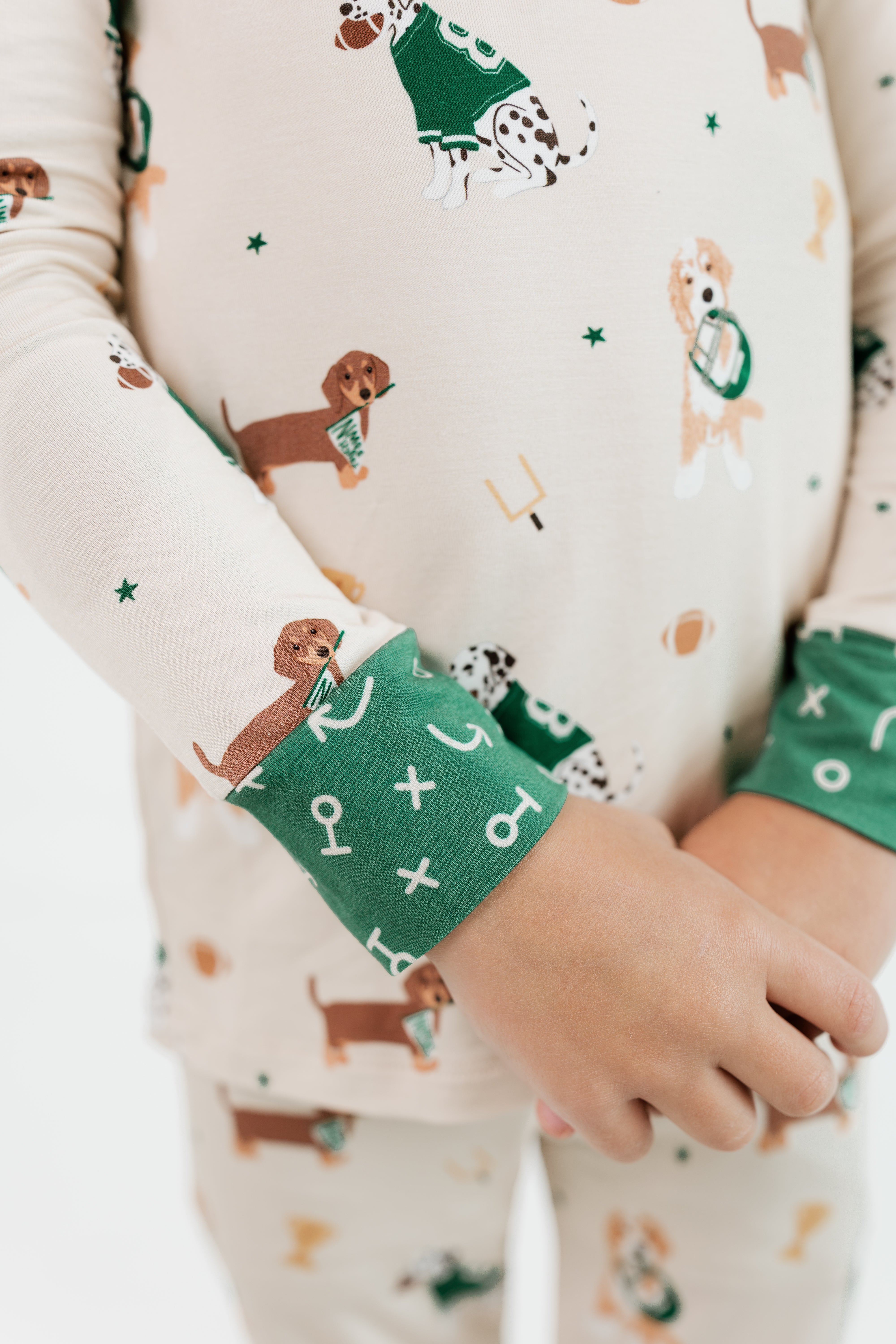 A close-up of a child wearing a long-sleeve pajama top with a cream base and a dog-themed print, featuring green cuffs with white symbols. The child's hands are clasped in front of the torso.