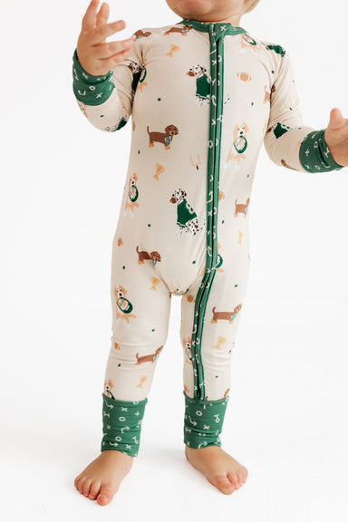A baby wearing a long-sleeve zip-front footless sleeper in cream with green cuffs and hem, printed with small dog illustrations, photographed against a plain white background.