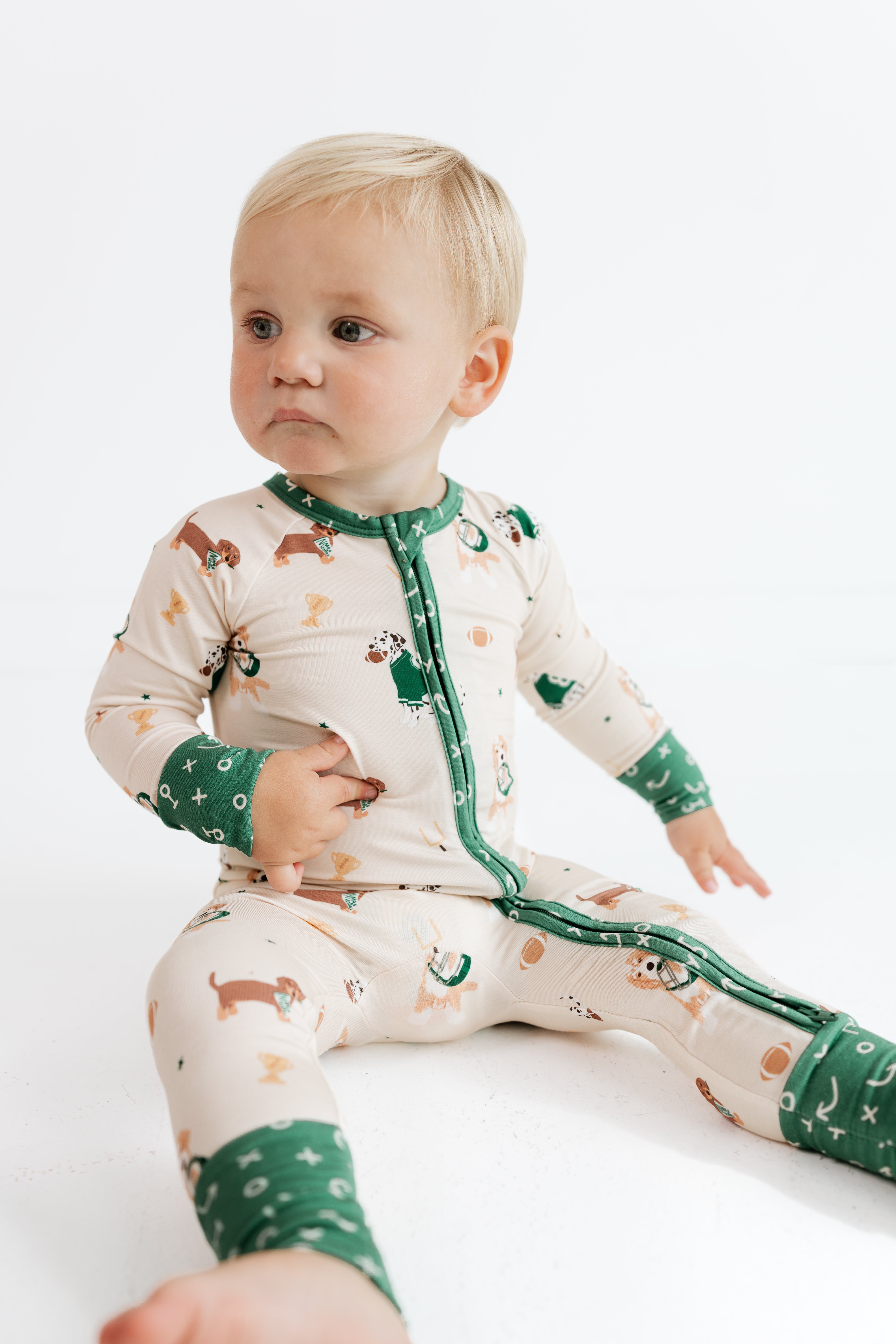 A baby sitting on a white studio floor wearing a cream-colored long-sleeve footed pajama with green cuffs and collar, featuring a dachshund and Christmas tree pattern.