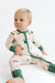 A baby sitting on a white studio floor wearing a cream-colored long-sleeve footed pajama with green cuffs and collar, featuring a dachshund and Christmas tree pattern.