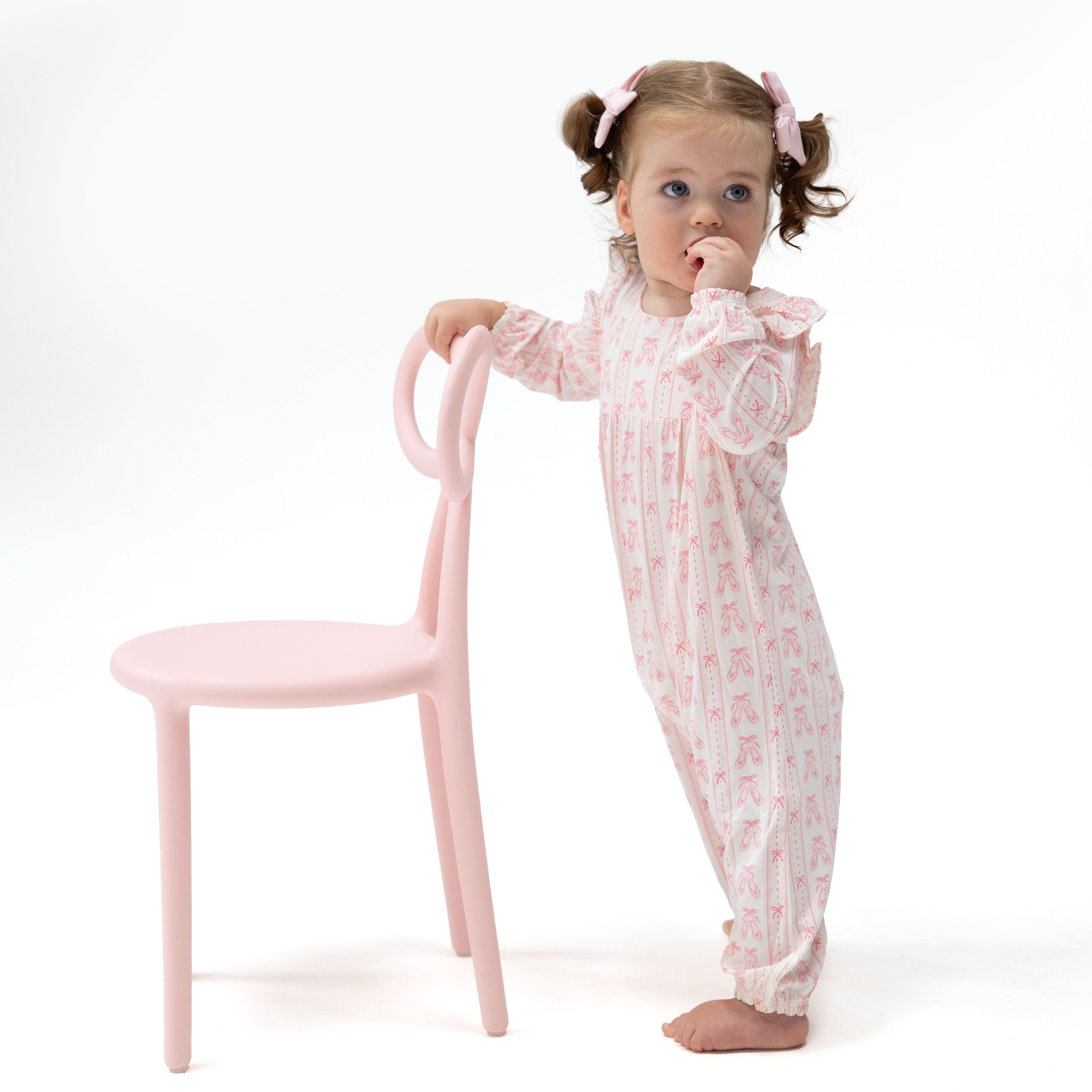 A toddler girl with pigtails stands barefoot next to a light pink chair, wearing a light pink and white long-sleeve footless pajama romper with a small repeating ballet slipper and bow pattern.
