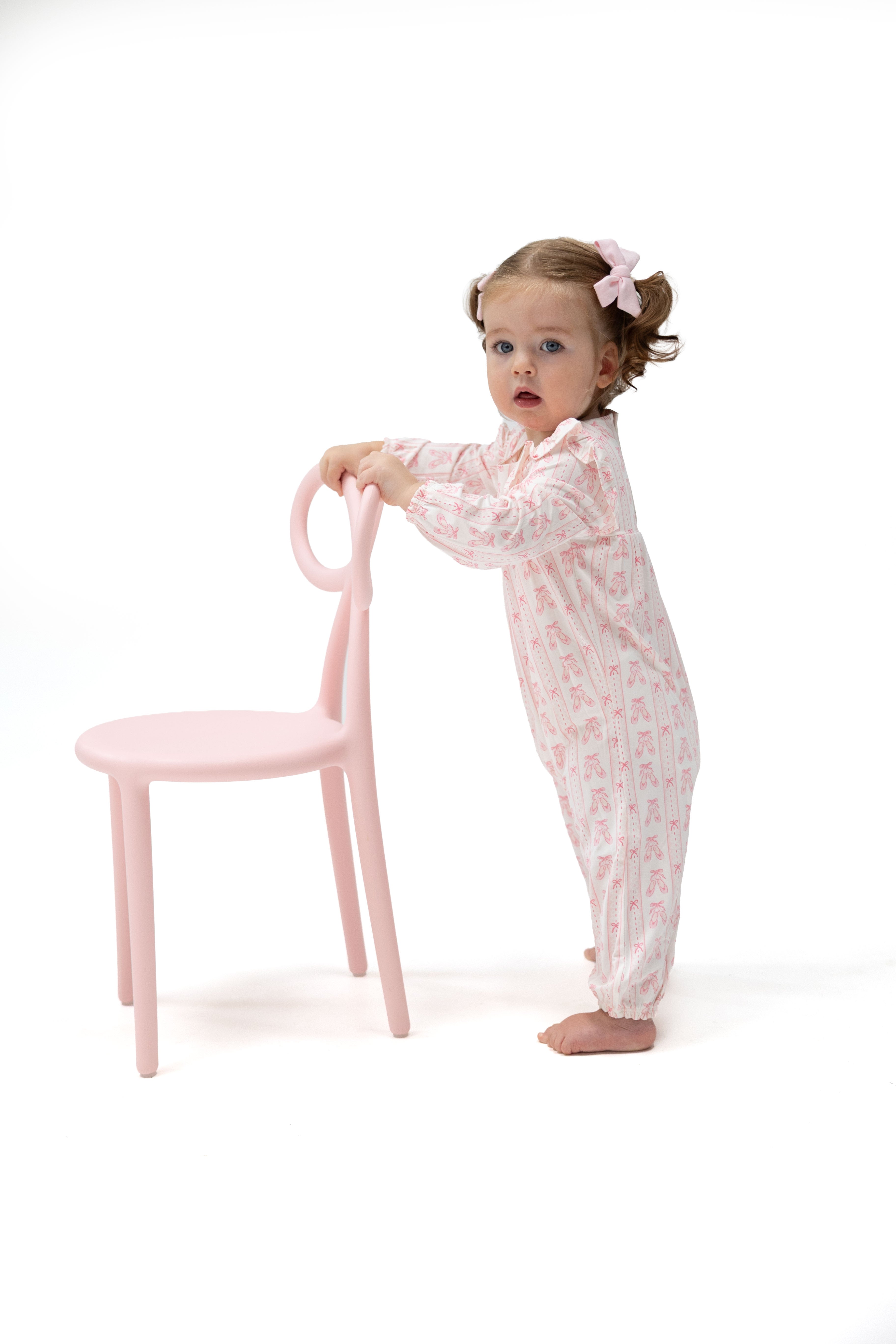 A toddler girl wearing a light pink patterned footed pajama romper stands barefoot next to a matching light pink chair, holding its backrest against a solid white background.