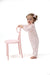 A toddler girl wearing a light pink patterned footed pajama romper stands barefoot next to a matching light pink chair, holding its backrest against a solid white background.