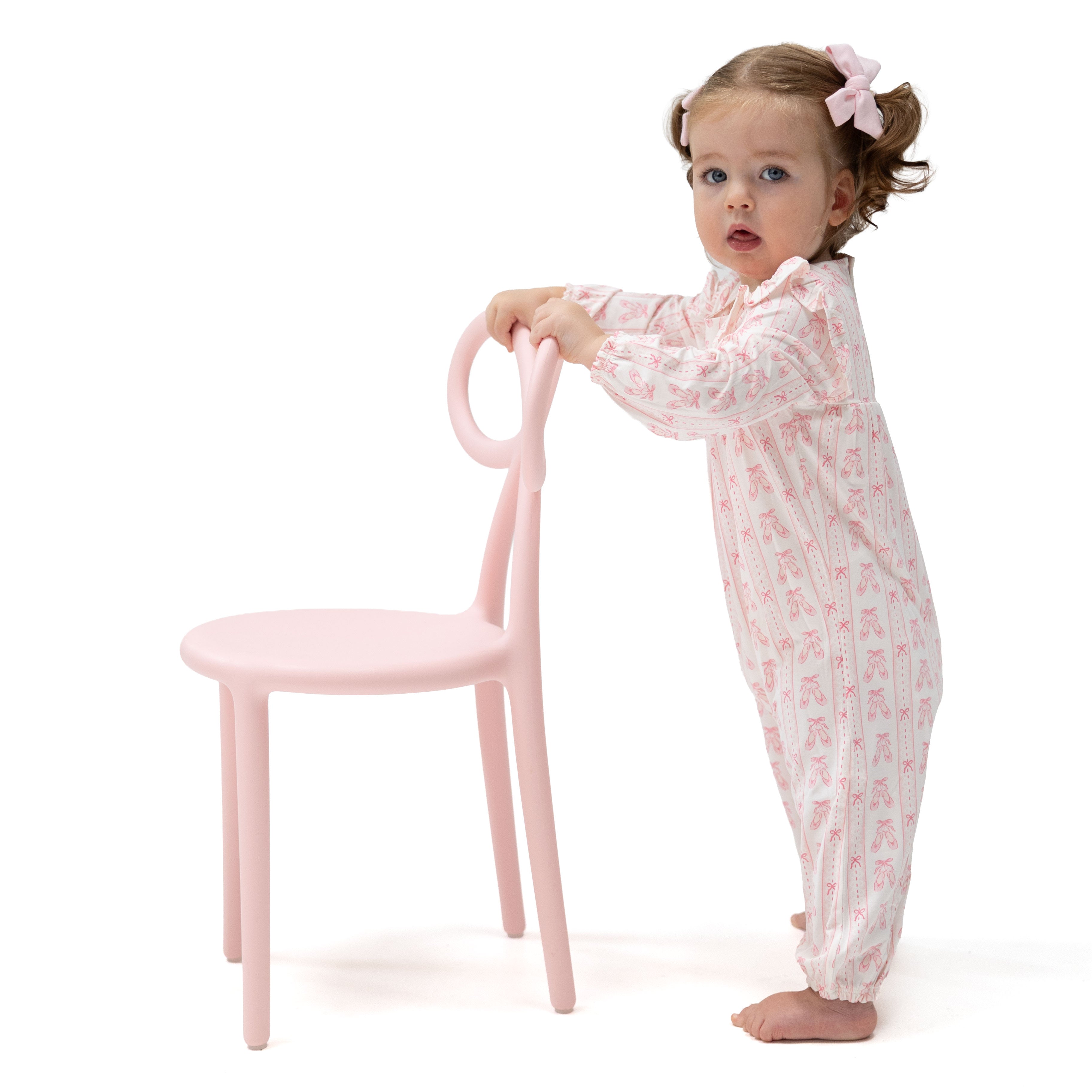 A toddler stands barefoot next to a light pink chair, wearing a light pink and white patterned footed pajama romper with a matching pink bow in her hair, photographed on a solid white background.