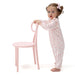 A toddler stands barefoot next to a light pink chair, wearing a light pink and white patterned footed pajama romper with a matching pink bow in her hair, photographed on a solid white background.