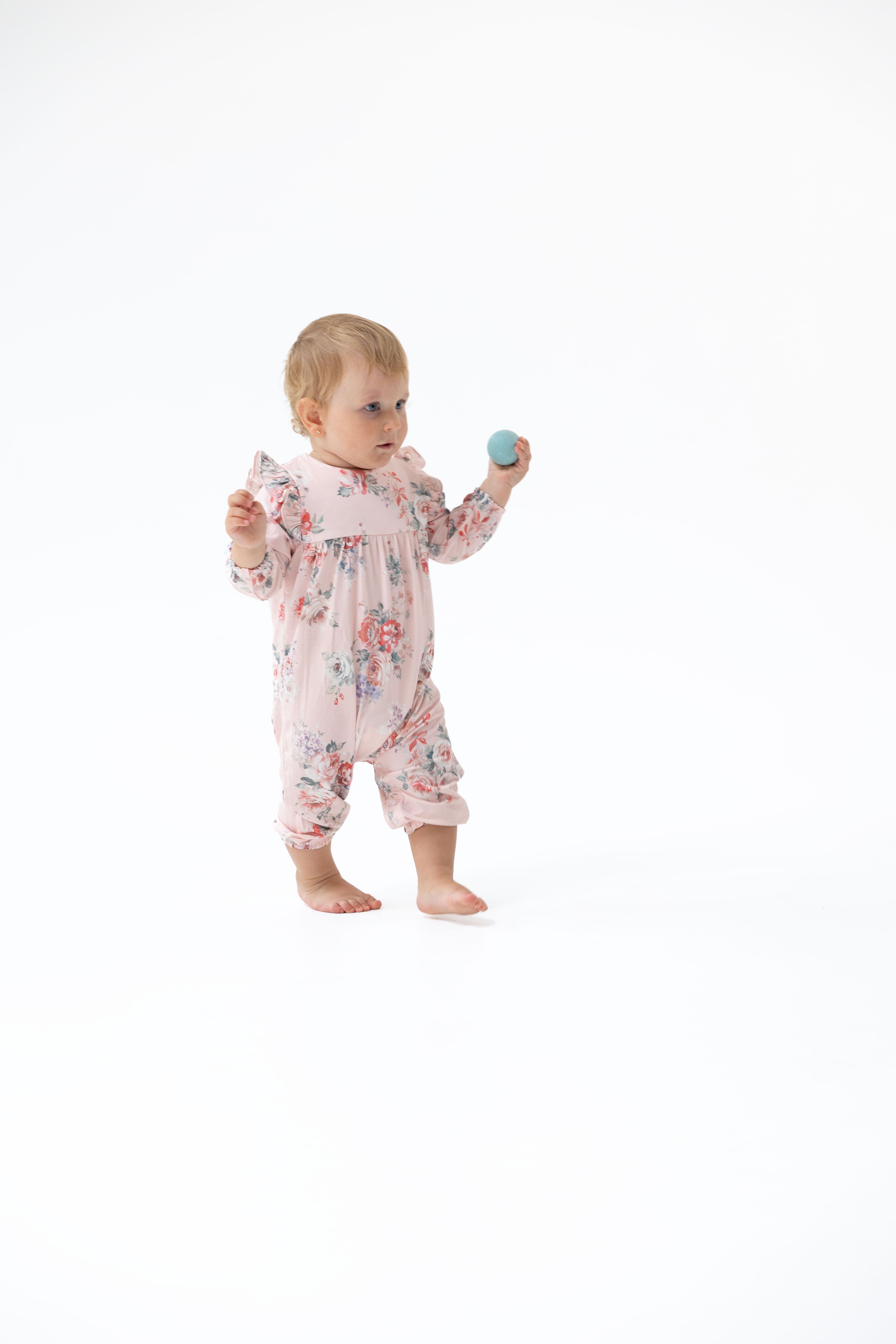 A baby standing barefoot on a white studio background, wearing a light pink floral long-sleeve romper and holding a small blue rattle.