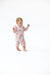 A baby standing barefoot on a white studio background, wearing a light pink floral long-sleeve romper and holding a small blue rattle.