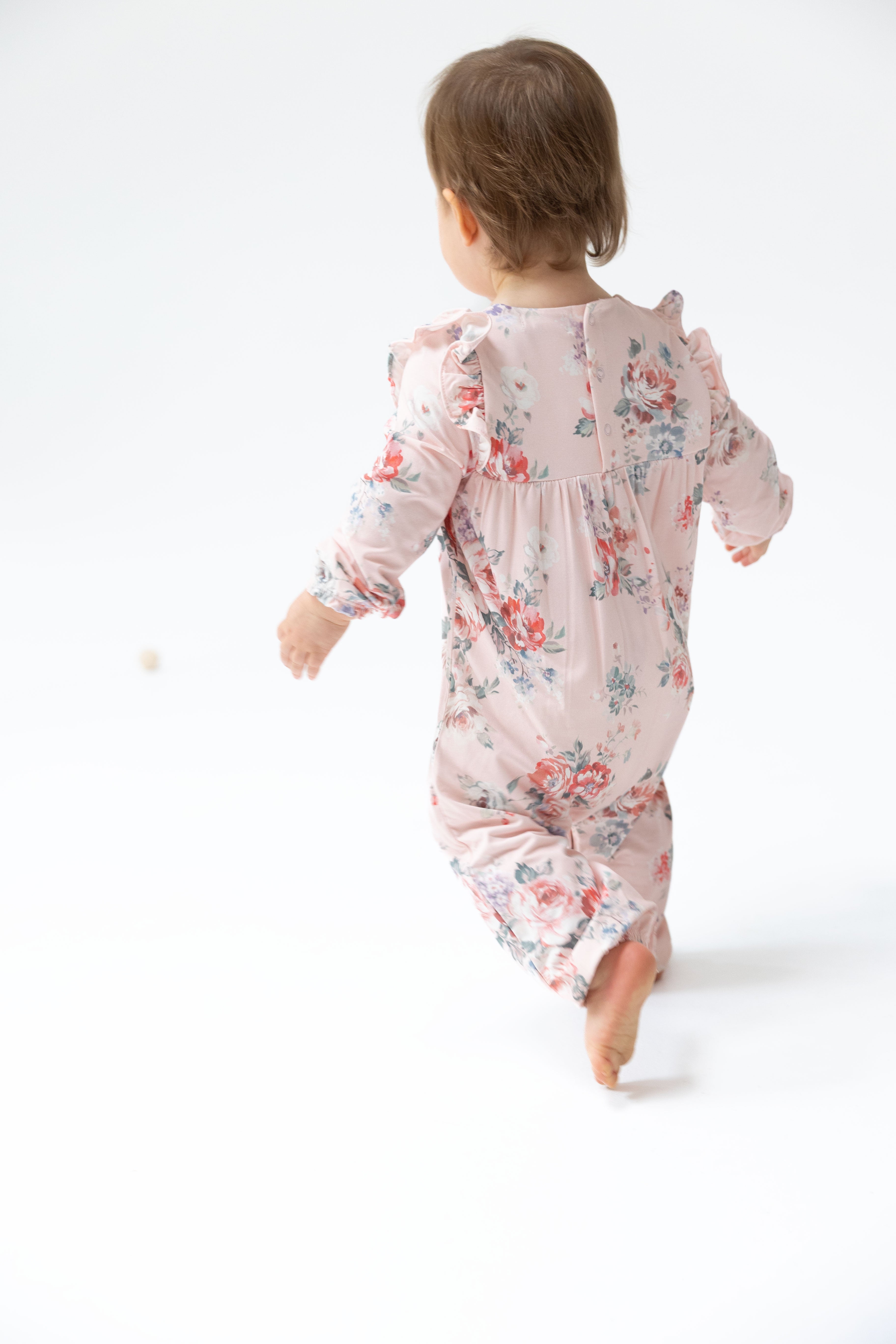 A baby wearing a light pink long-sleeve footless sleeper with a red and blue floral print, shown from behind as they walk barefoot on a white background.