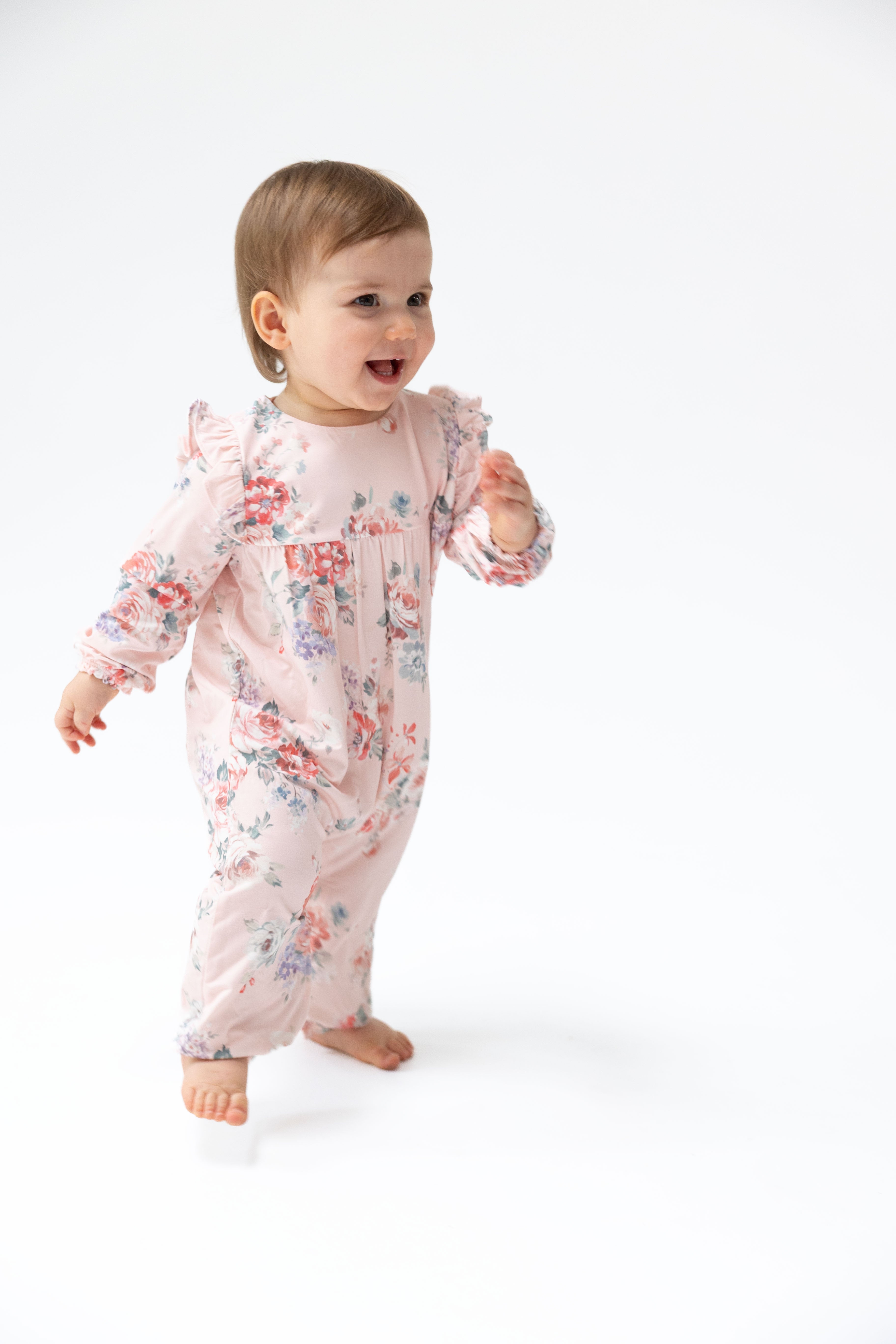 A baby girl wearing a light pink long-sleeve footless romper with a red and blue floral print, walking barefoot on a white studio background.