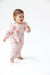 A baby girl wearing a light pink long-sleeve footless romper with a red and blue floral print, walking barefoot on a white studio background.