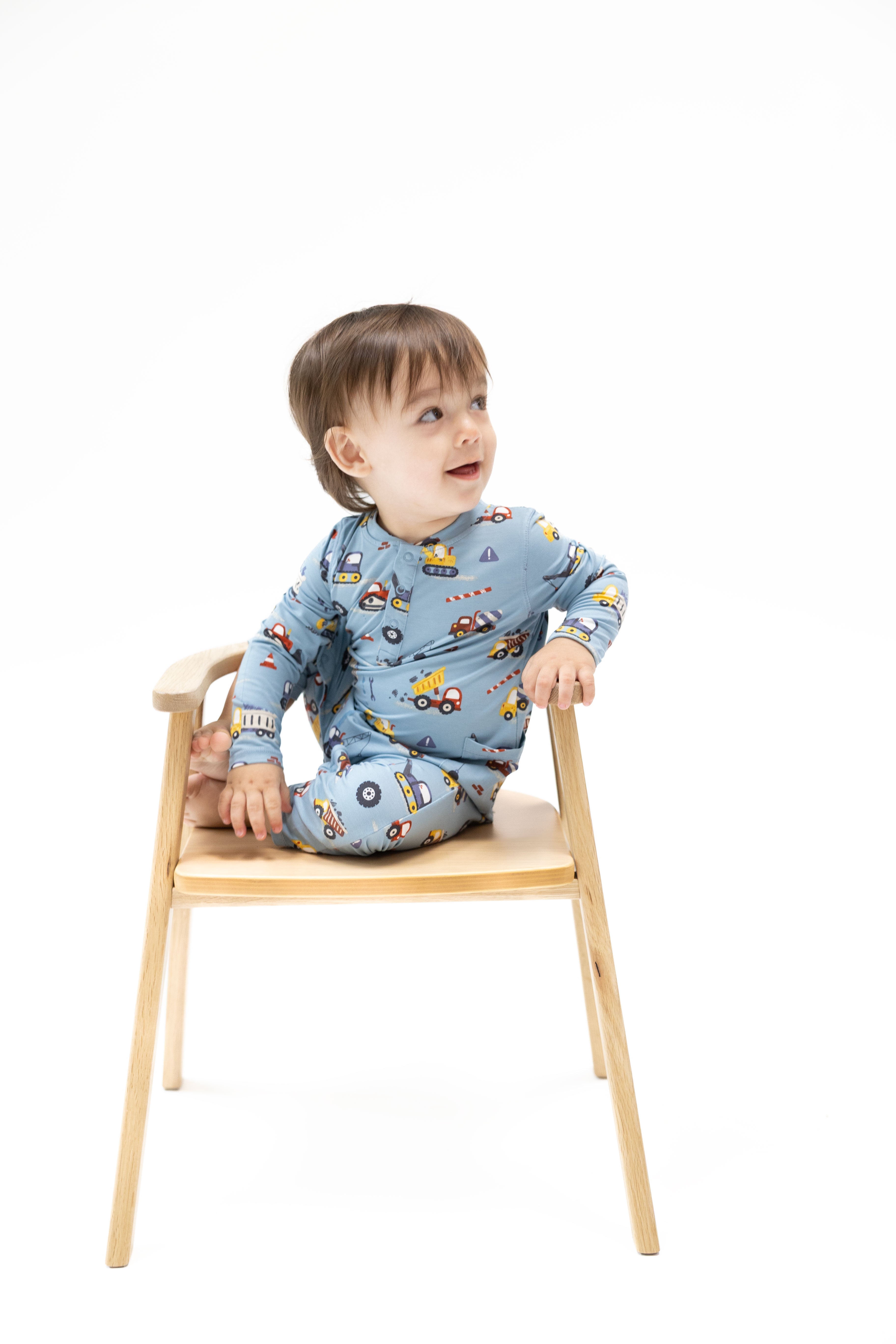 A toddler sitting on a light wood chair against a white background, wearing a light blue long-sleeve pajama set with a colorful construction vehicle print.
