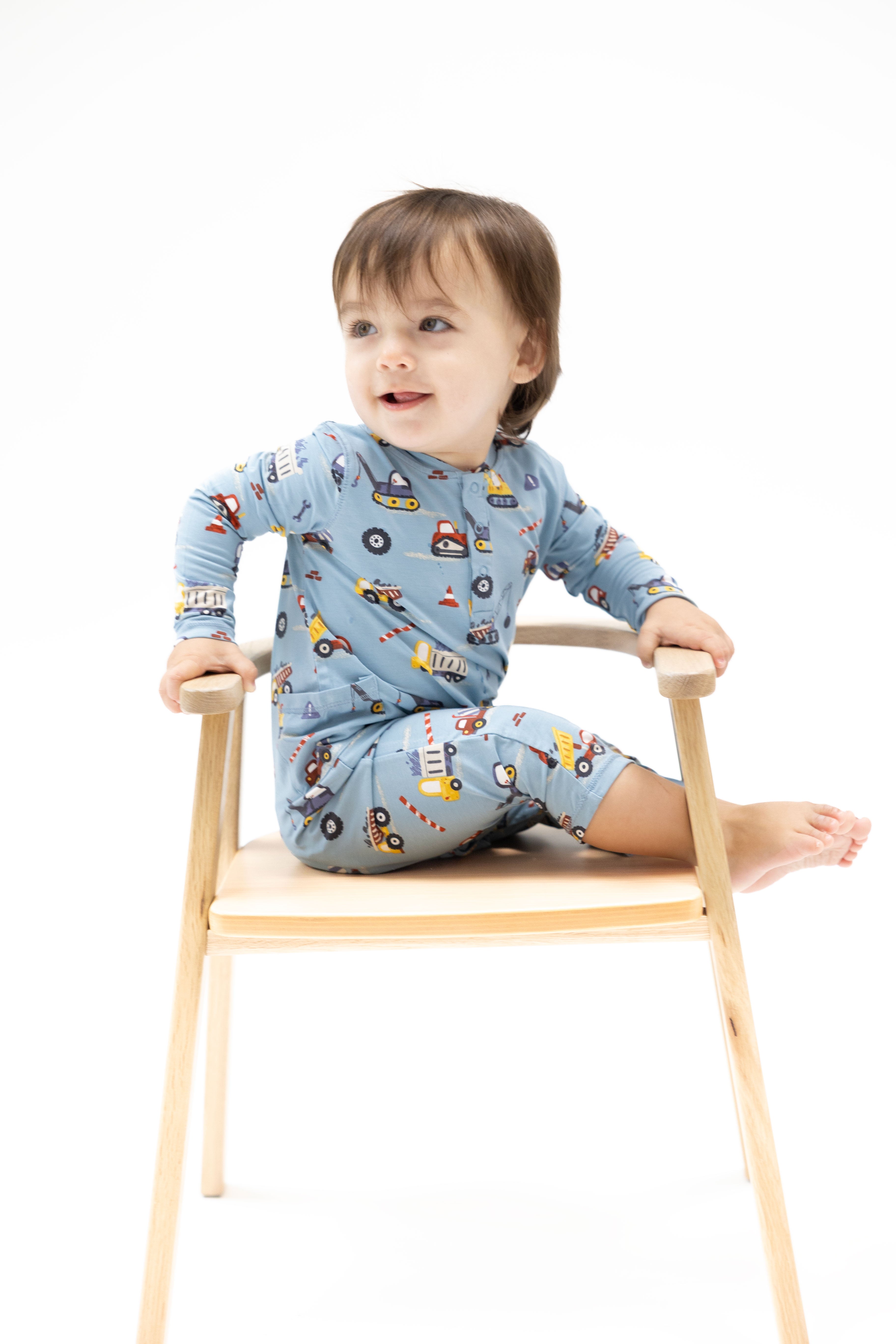 A toddler wearing a light blue long-sleeve pajama set with a colorful construction vehicle print, sitting on a light wood chair against a white background.