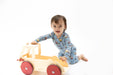 A toddler in light blue patterned pajamas pushes a wooden toy truck with red wheels on a seamless white background.