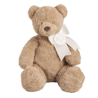 Beige teddy bear with a white bow and mongram tag on a white background