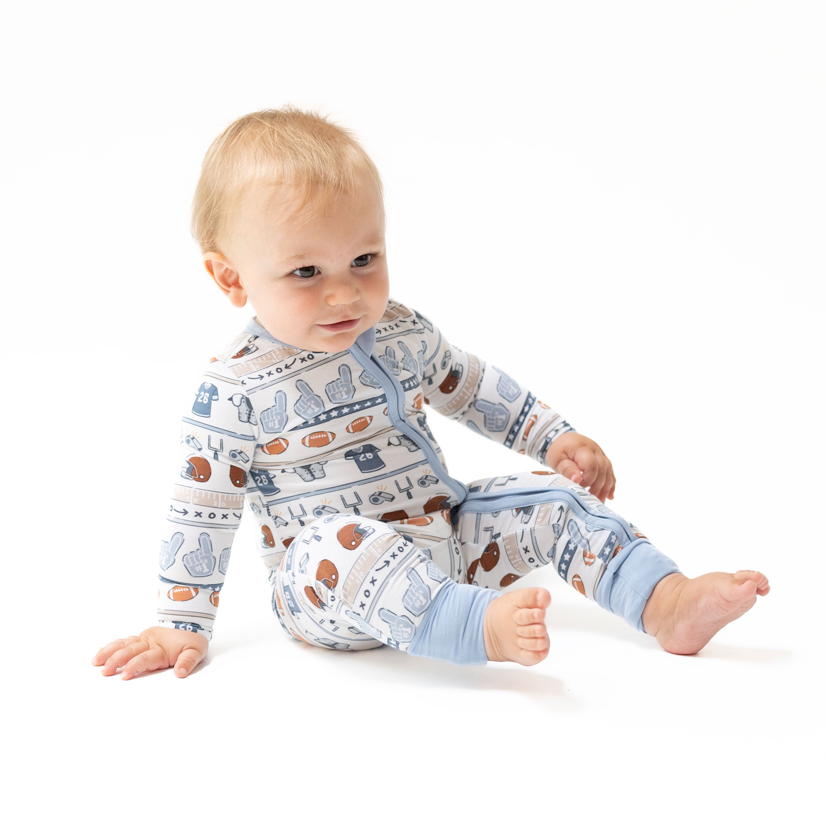 A baby sitting on a white studio background wearing a long-sleeve footless pajama romper with a light blue and white patterned design featuring football equipment.