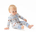 A baby sitting on a white studio background wearing a long-sleeve footless pajama romper with a light blue and white patterned design featuring football equipment.