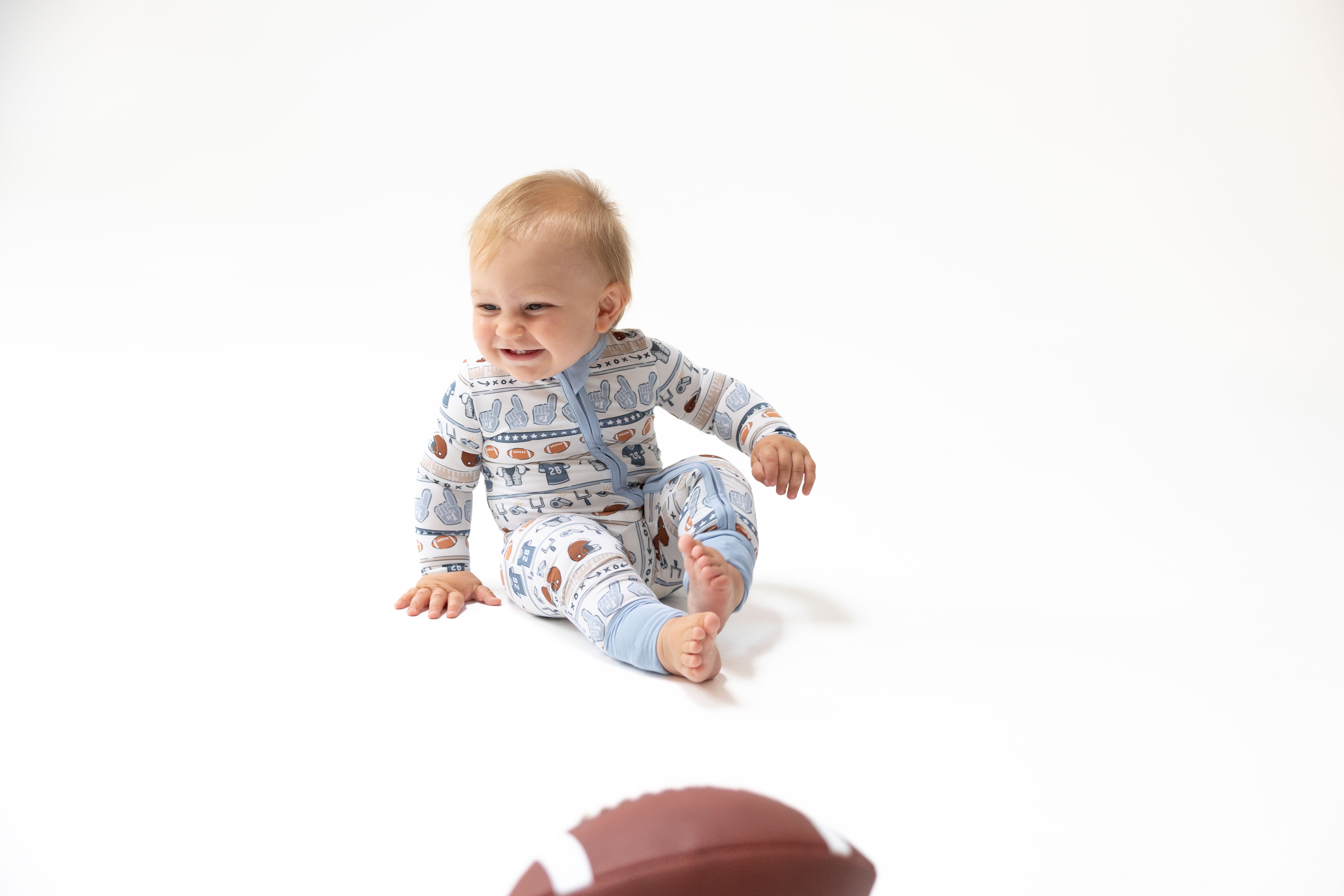 A baby sitting on a white studio floor wearing a light blue and white patterned long-sleeve footed sleeper, with a brown football blurred in the foreground.