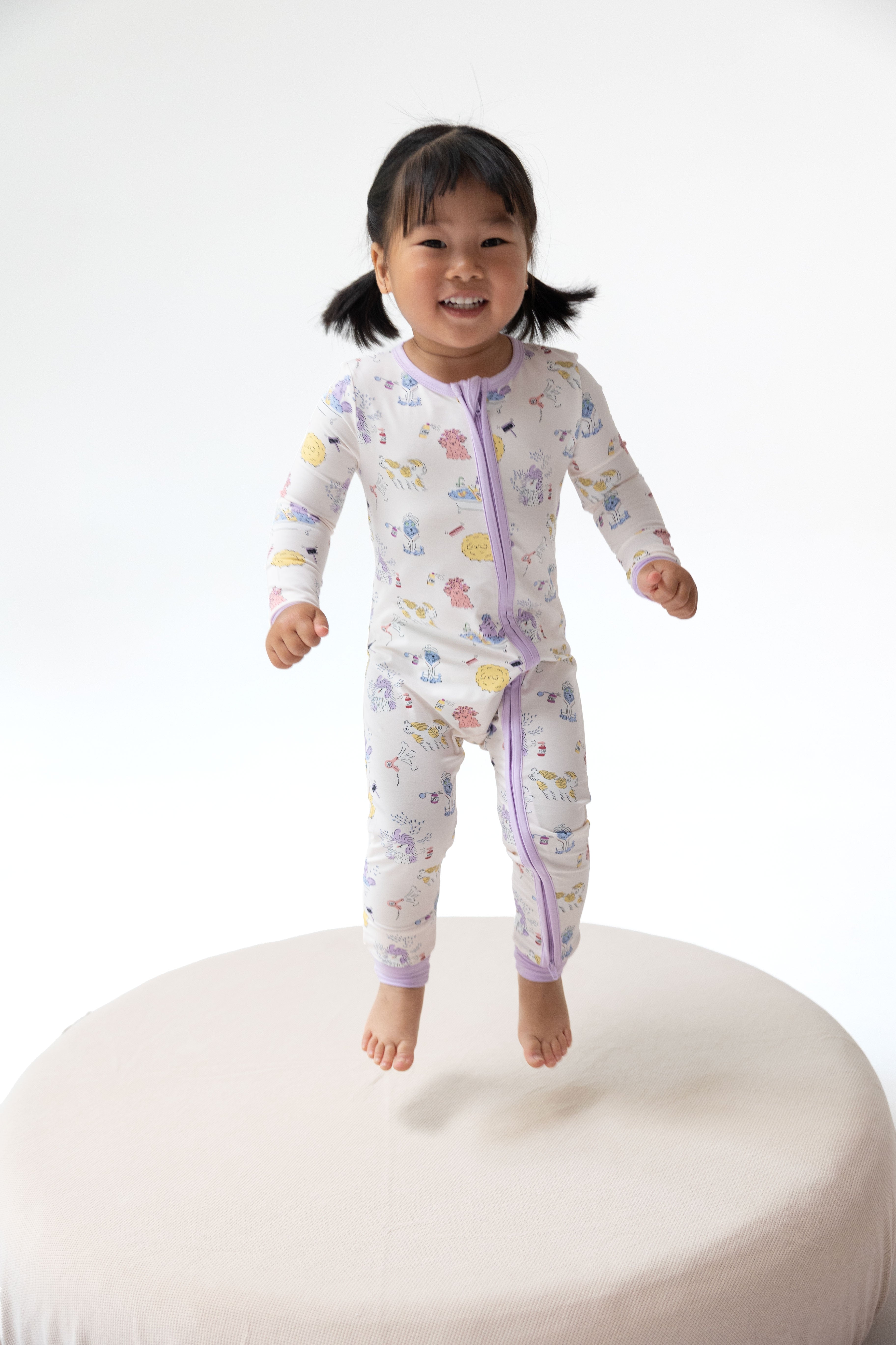 A smiling toddler girl jumps barefoot on a round white platform, wearing a long-sleeve zip-front sleeper with a light lavender base and a colorful print of cartoon animals and planets.