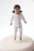 A smiling toddler girl jumps barefoot on a round white platform, wearing a long-sleeve zip-front sleeper with a light lavender base and a colorful print of cartoon animals and planets.