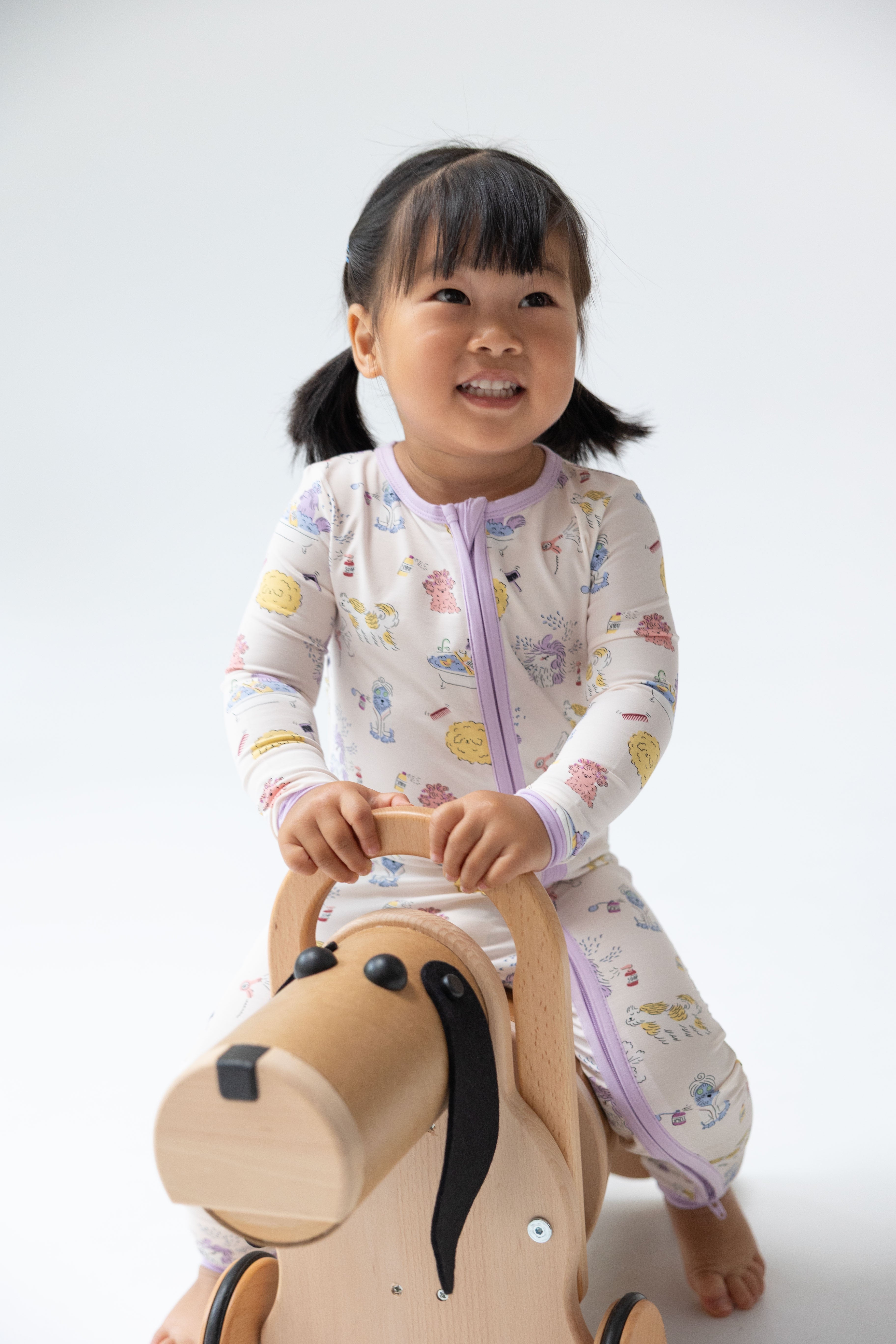 A smiling young child wearing a light cream zip-up sleeper with a pastel multicolor animal print, sitting on a wooden horse toy against a clean white studio background.