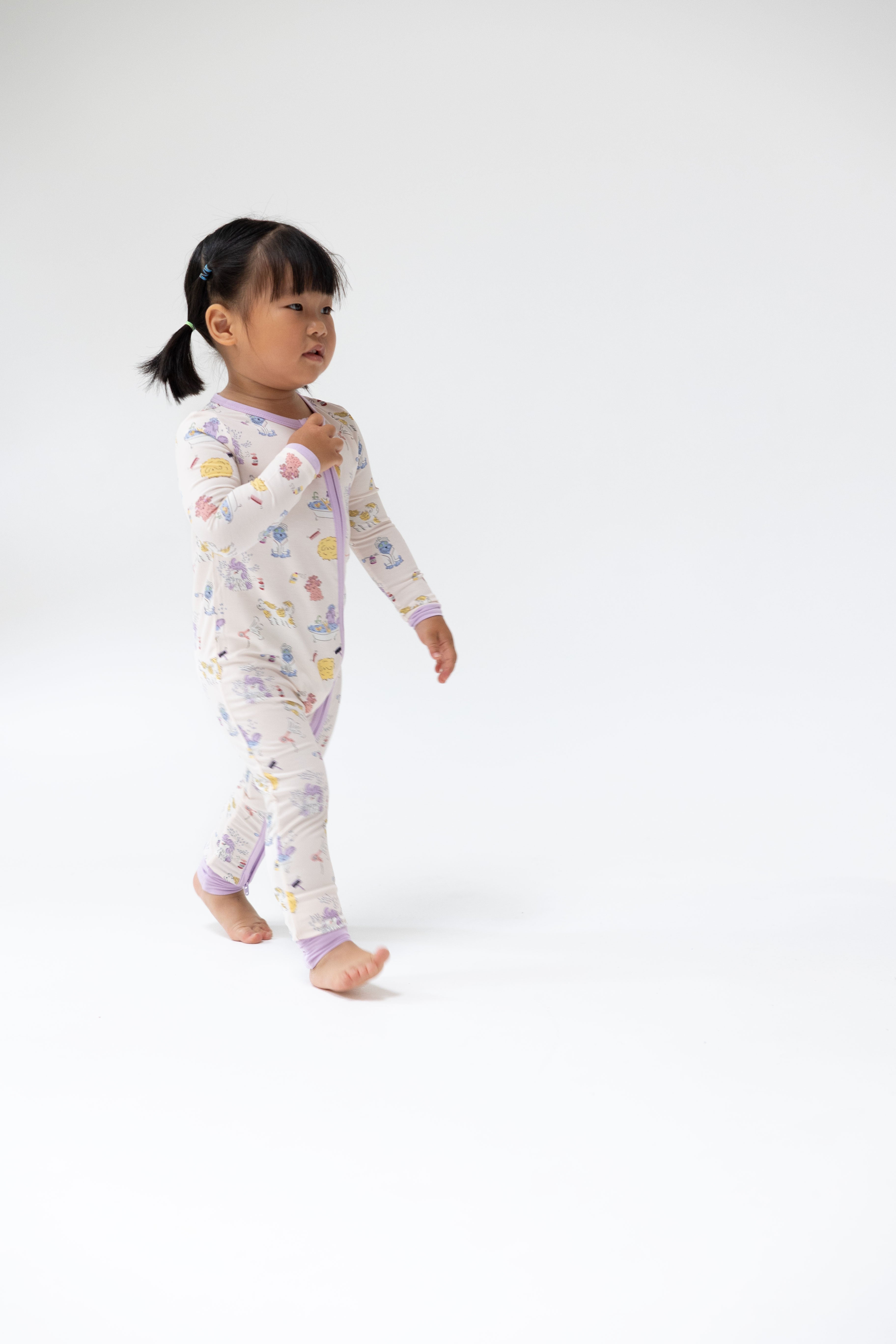 A toddler girl walking barefoot in a white long-sleeve pajama romper with a colorful cartoon pattern, photographed against a seamless white studio background.