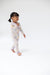 A toddler girl walking barefoot in a white long-sleeve pajama romper with a colorful cartoon pattern, photographed against a seamless white studio background.