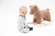 A baby wearing a white and navy blue pinstripe pajama set sits on a white studio floor next to a brown plush rocking horse toy.