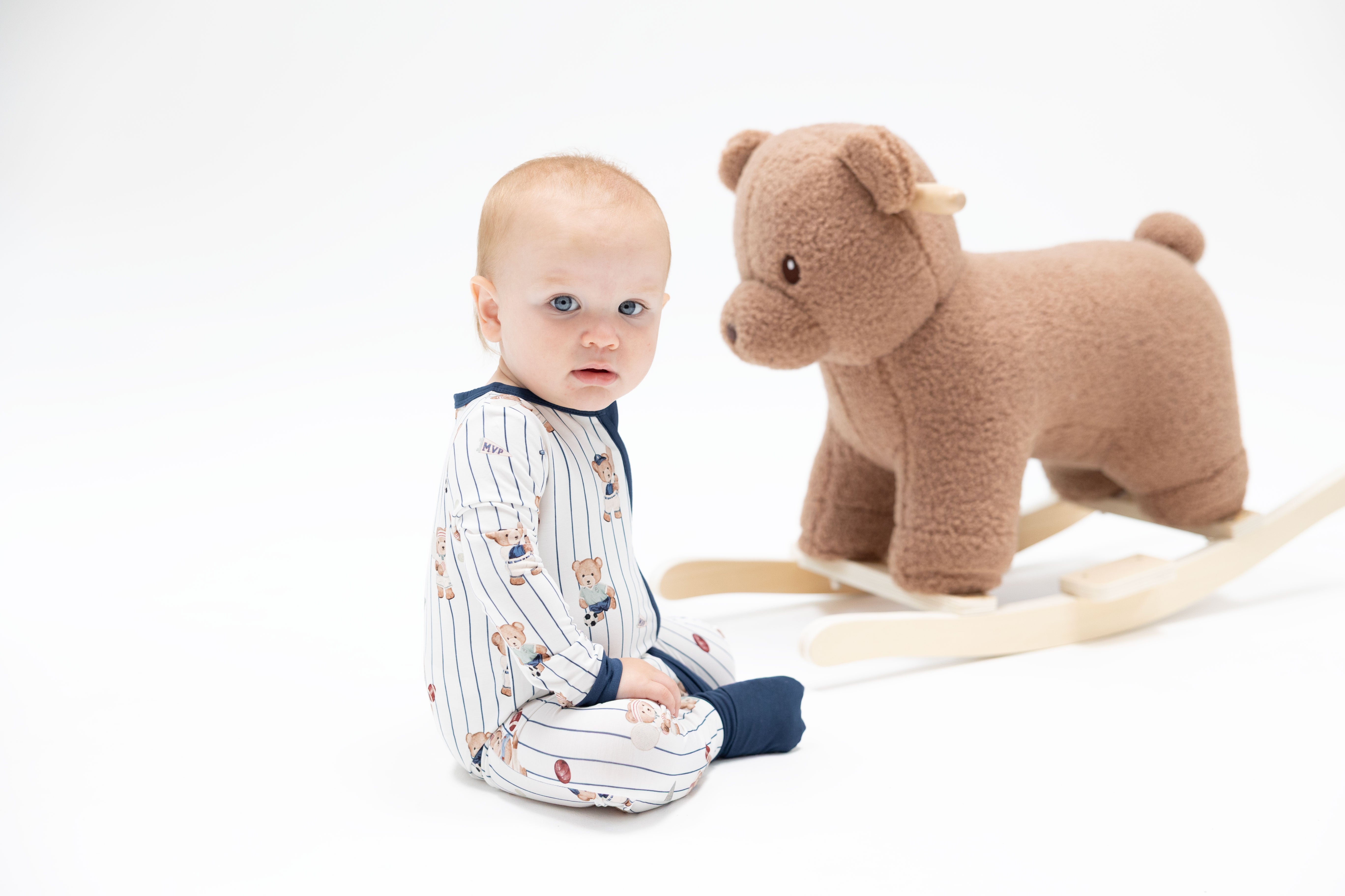 A baby wearing a white and navy blue pinstripe pajama set sits on a white studio floor next to a brown plush rocking horse toy.