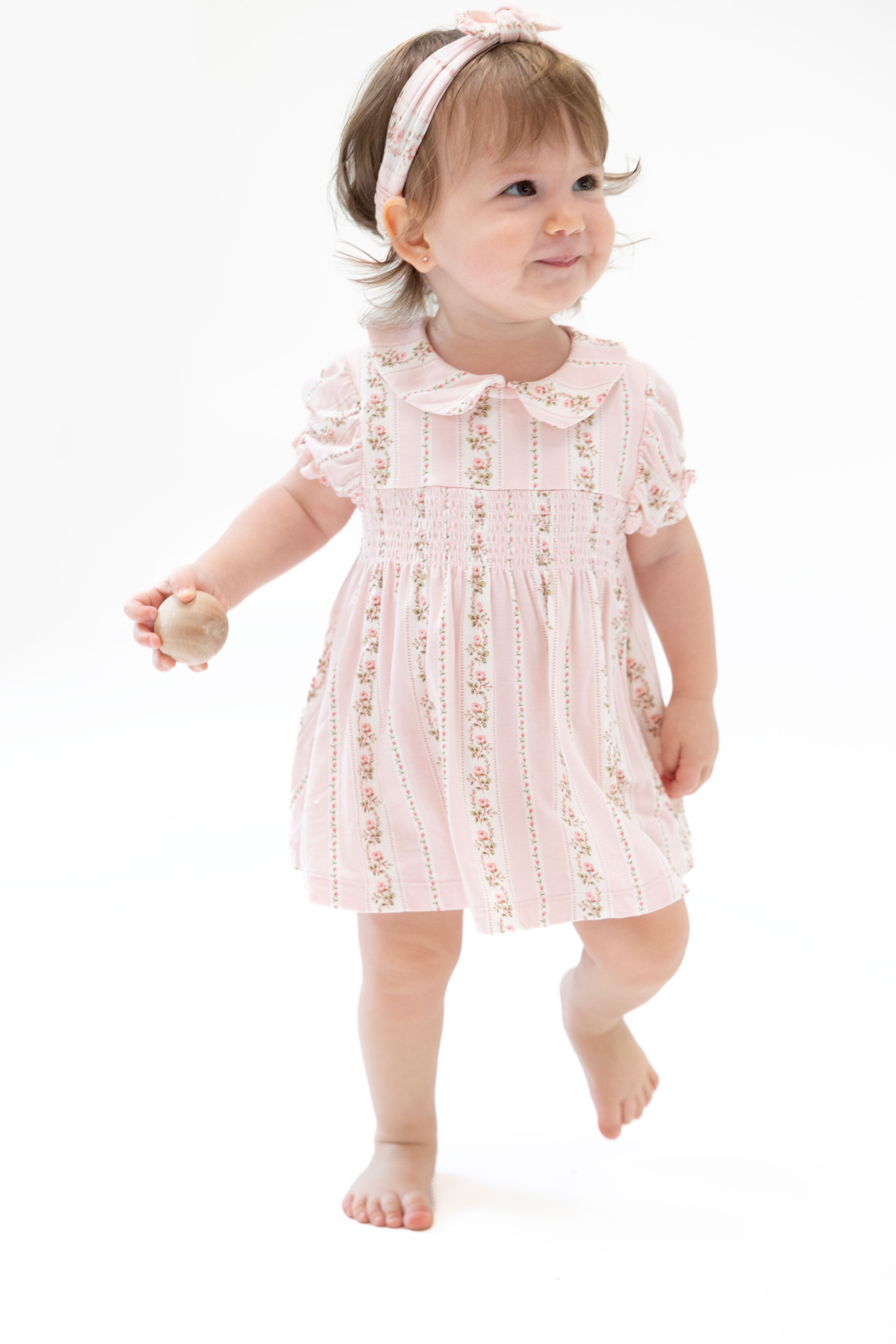 A toddler girl walking barefoot on a white studio background, wearing a light pink short-sleeve dress with vertical floral prints and a matching pink headband.