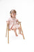 A toddler girl wearing a light pink embroidered dress and matching jacket sits barefoot on a small wooden chair against a solid white background.