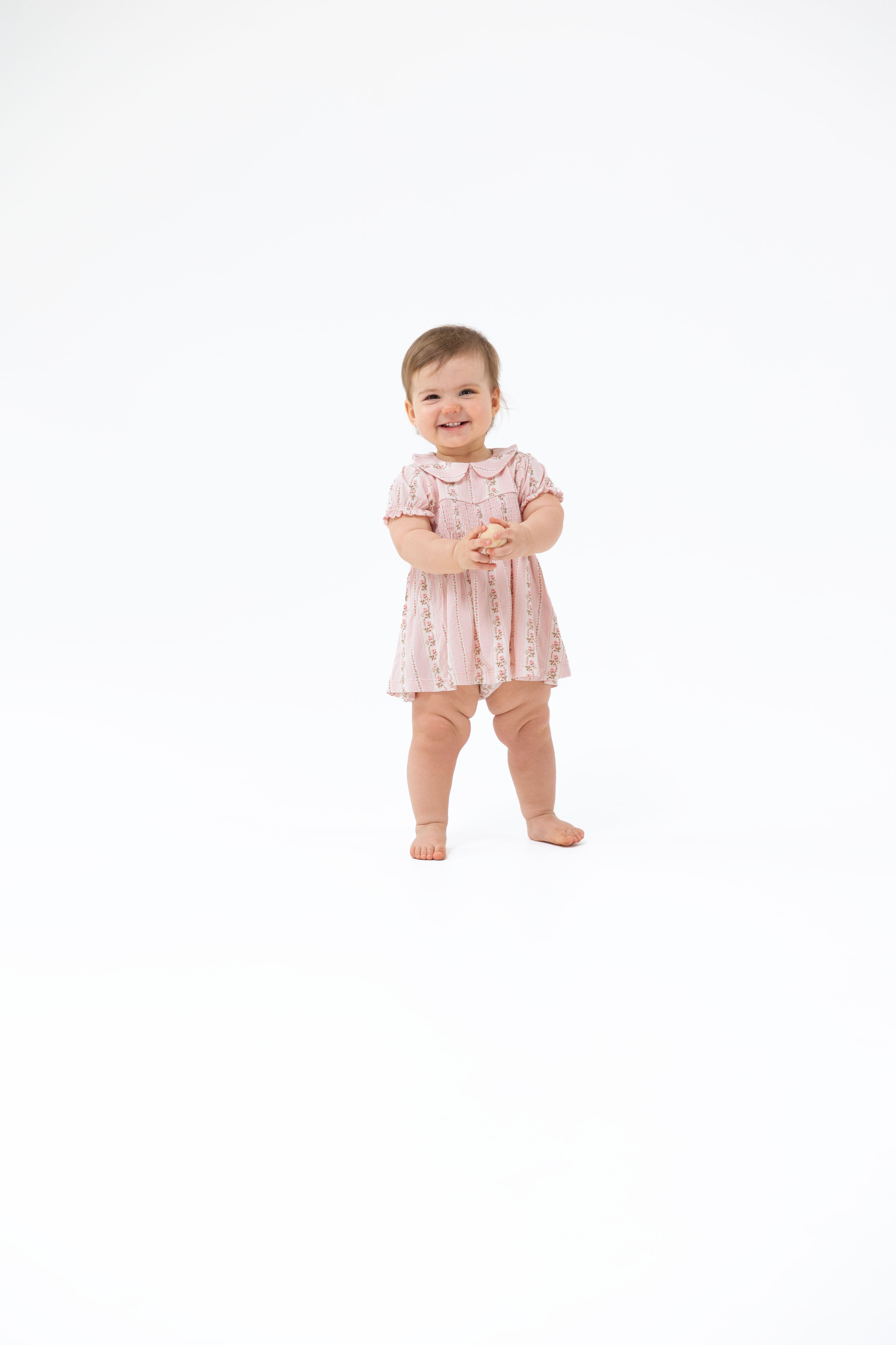 A smiling toddler standing on a seamless white studio background, wearing a light pink patterned dress with short sleeves and a Peter Pan collar, paired with matching pink leggings.