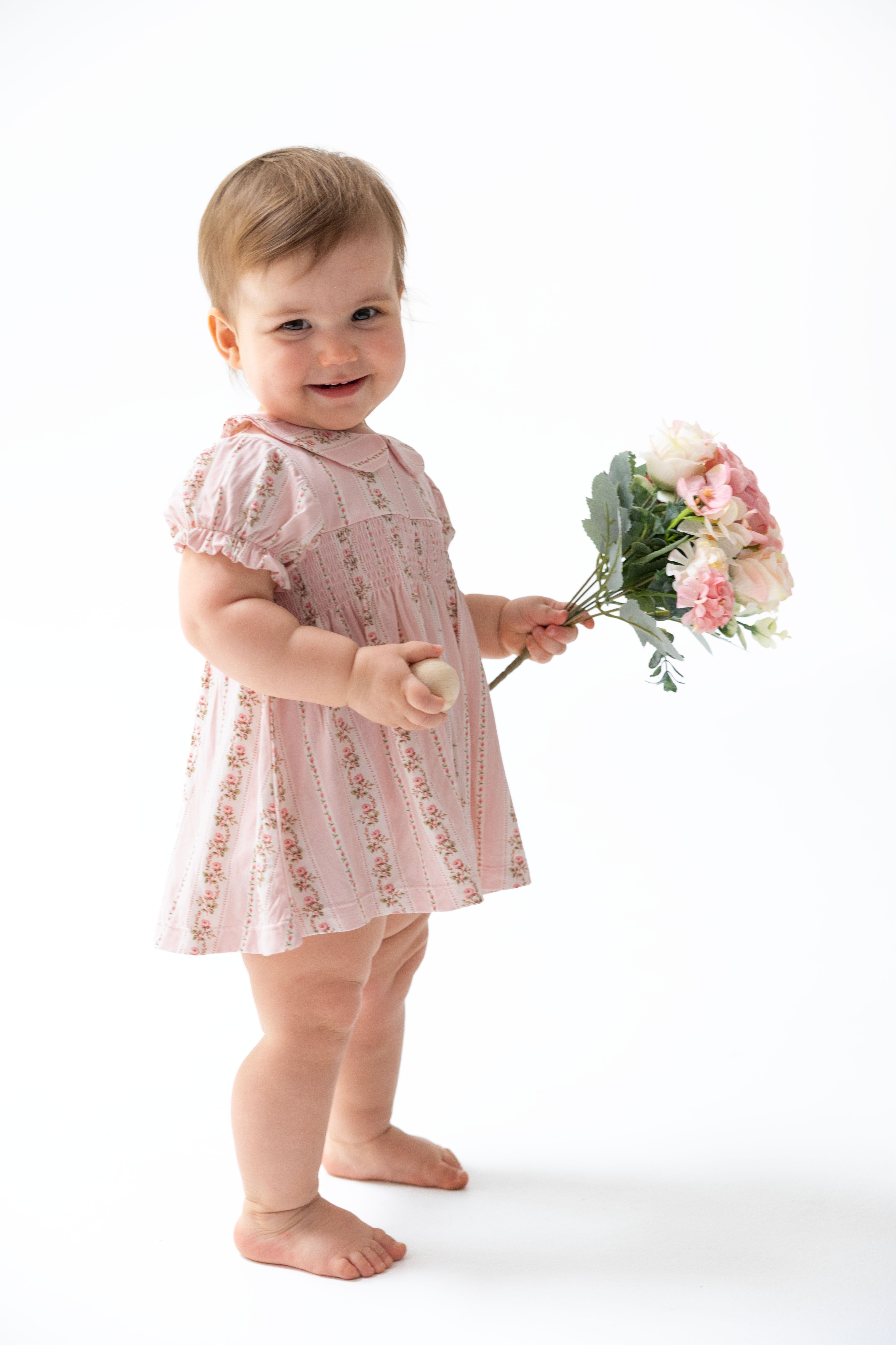 A smiling toddler standing barefoot on a white studio background, wearing a light pink short-sleeve dress with a subtle floral print and holding a bouquet of pink and white flowers.