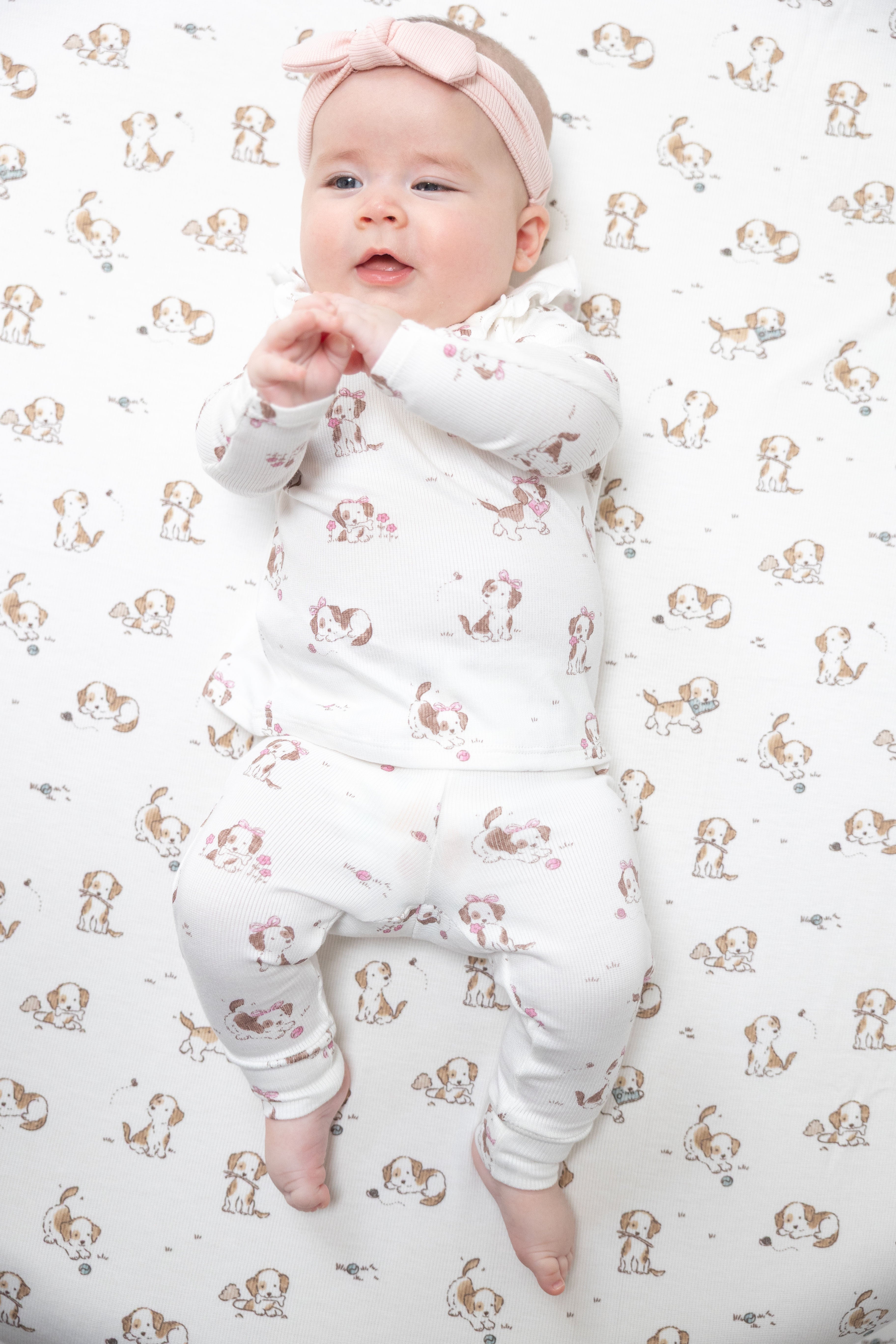 A baby lying on their back wearing a white long-sleeve ruffled top and pant set with a pink and brown dog pattern, along with a matching pink bow headband, on a matching patterned blanket.
