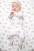 A baby lying on their back wearing a white long-sleeve ruffled top and pant set with a pink and brown dog pattern, along with a matching pink bow headband, on a matching patterned blanket.