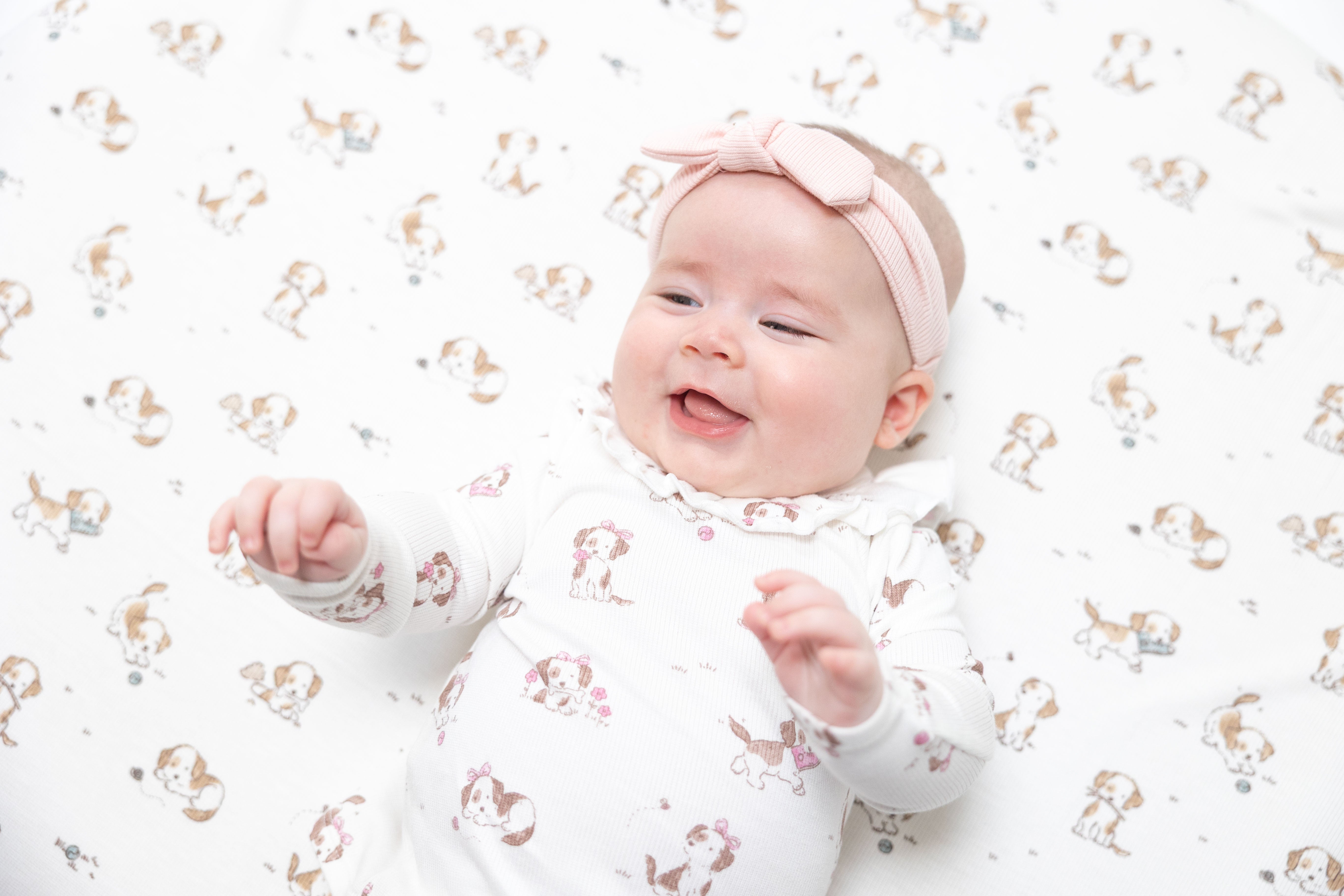 A smiling baby lying on a white blanket with a repeating dog print, wearing a white long-sleeve top and a pink bow headband.