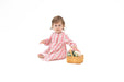 A toddler girl sits on a white studio background wearing a pink and white patterned long-sleeve pajama. She is holding a small woven basket with flowers.