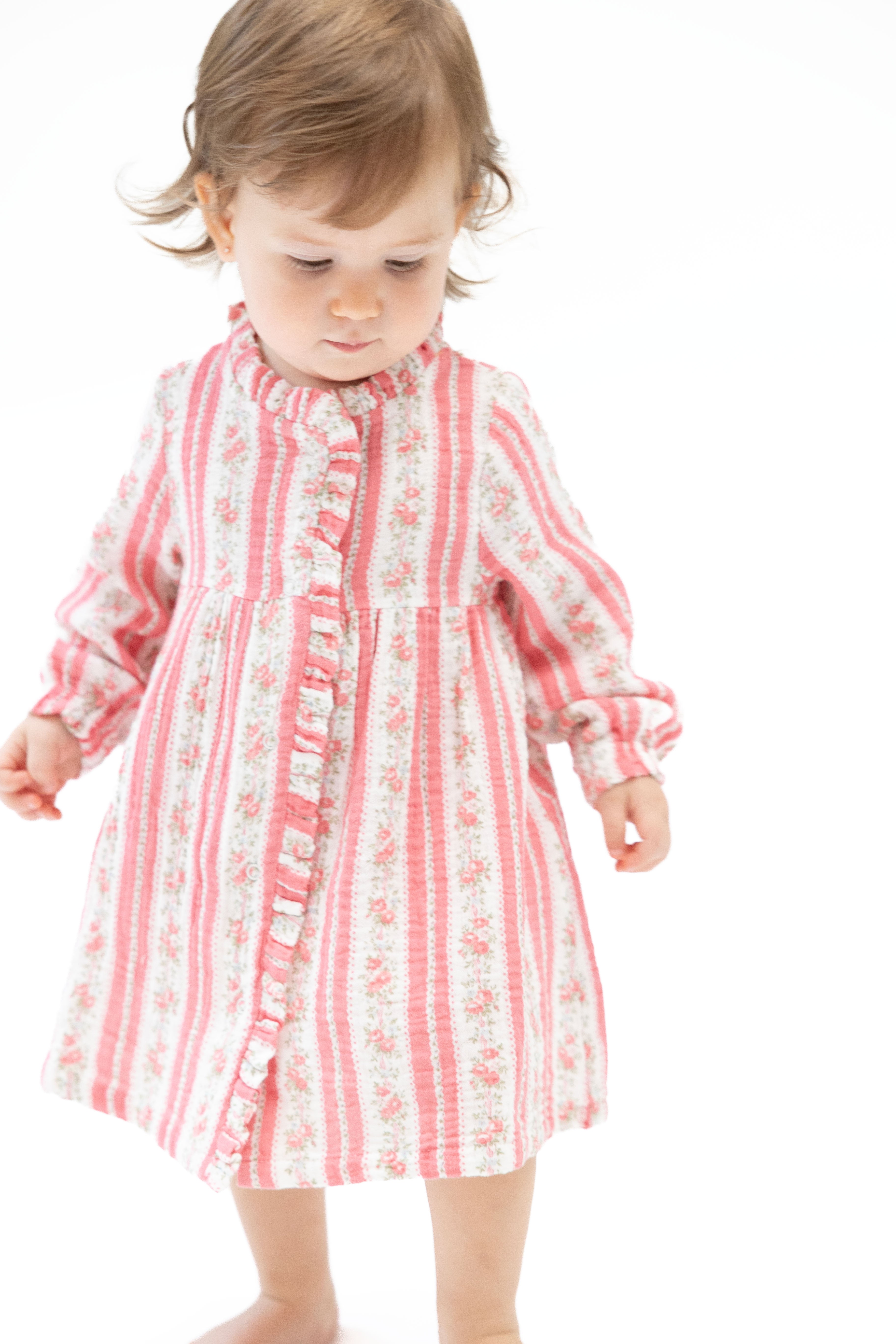 A toddler girl wearing a pink and white striped long-sleeve dress with a ruffled collar and button front, standing against a plain white background.