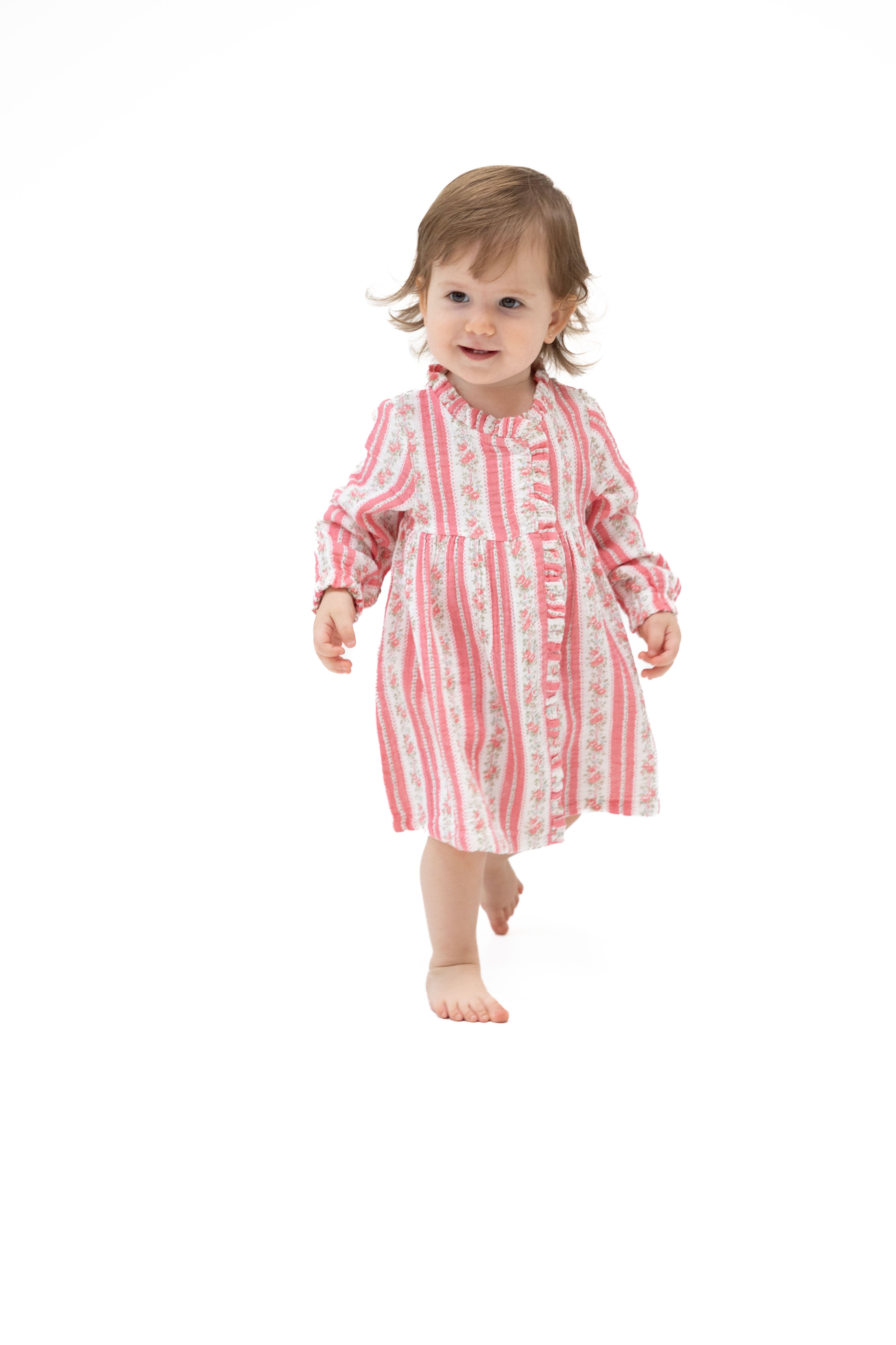 A toddler girl walking barefoot on a seamless white background, wearing a pink and white vertically striped long-sleeve nightgown with a floral pattern.