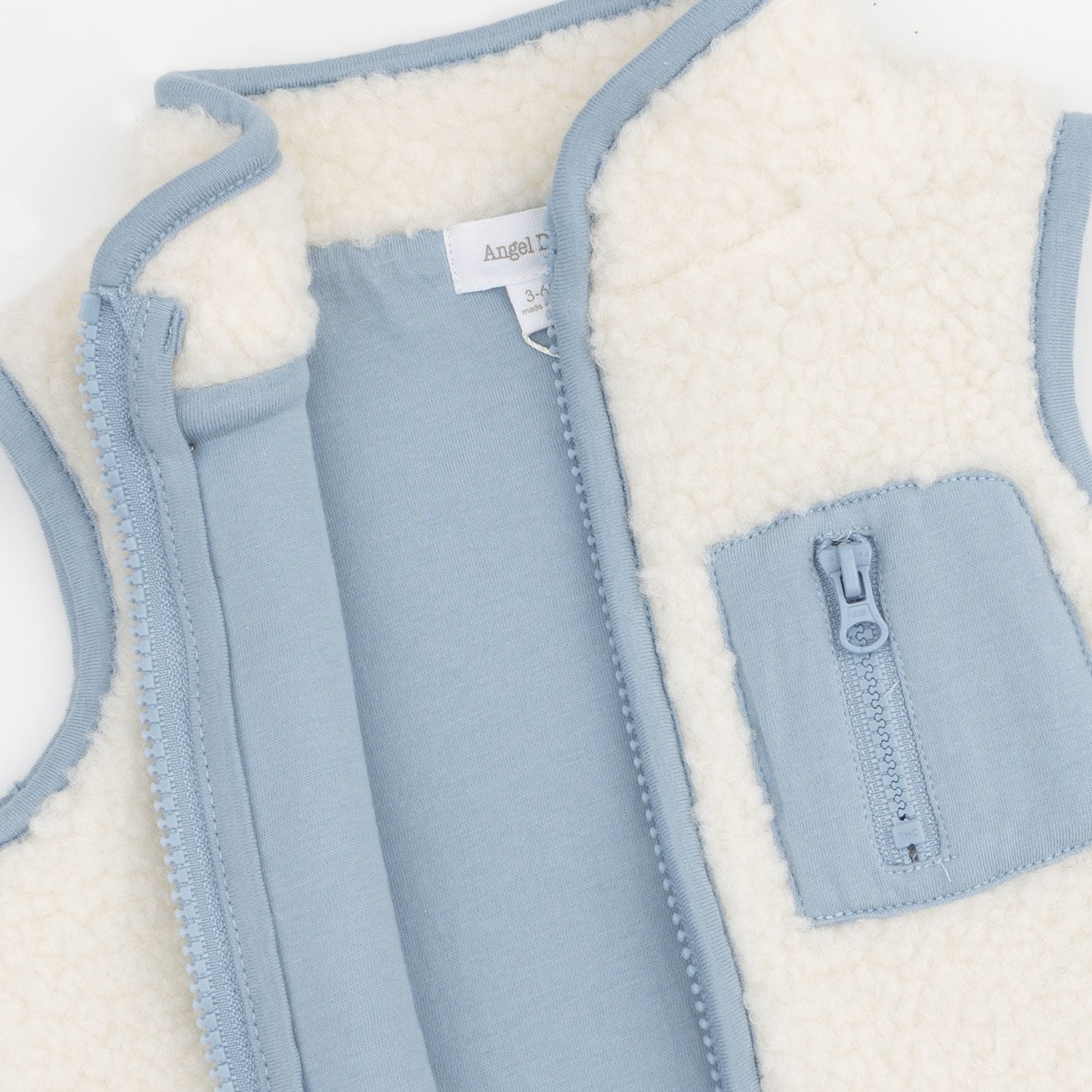 A close-up of a children's zip-front vest with a soft cream sherpa lining and light blue fabric exterior. A front pocket with a matching zipper is visible.