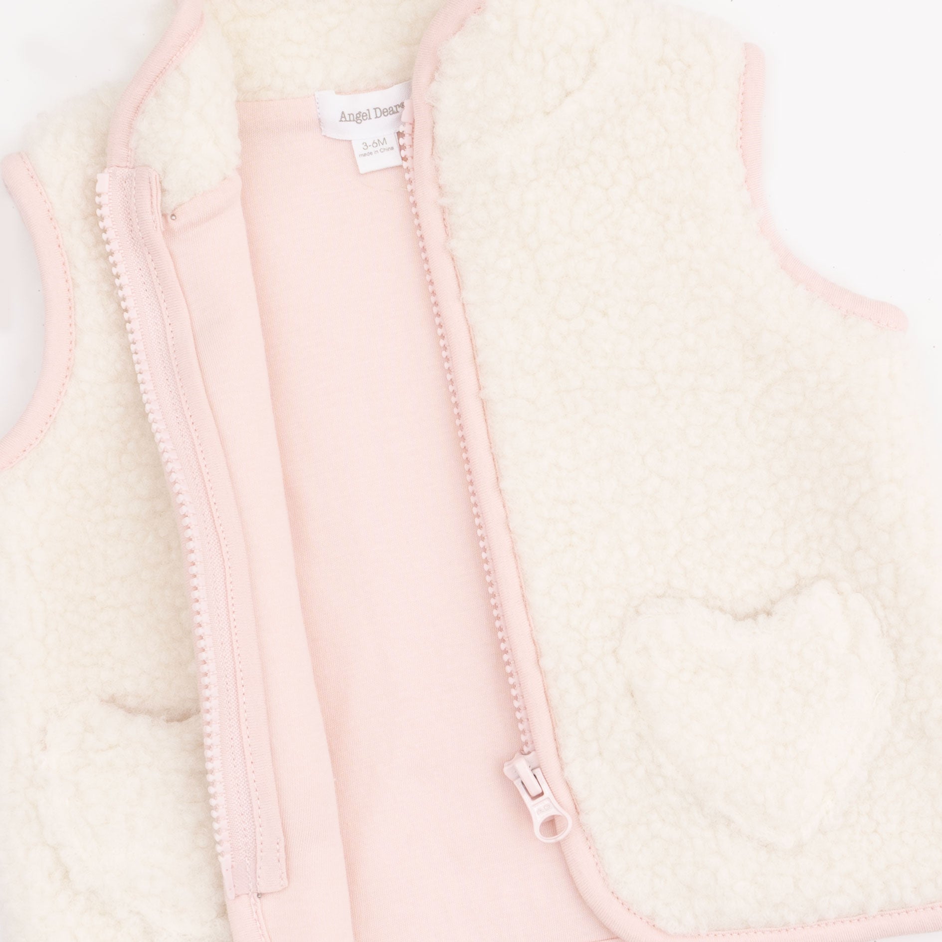A close-up of a children's zip-front vest with a soft cream sherpa fleece exterior, light pink lining and trim, and a heart-shaped patch on the front pocket.