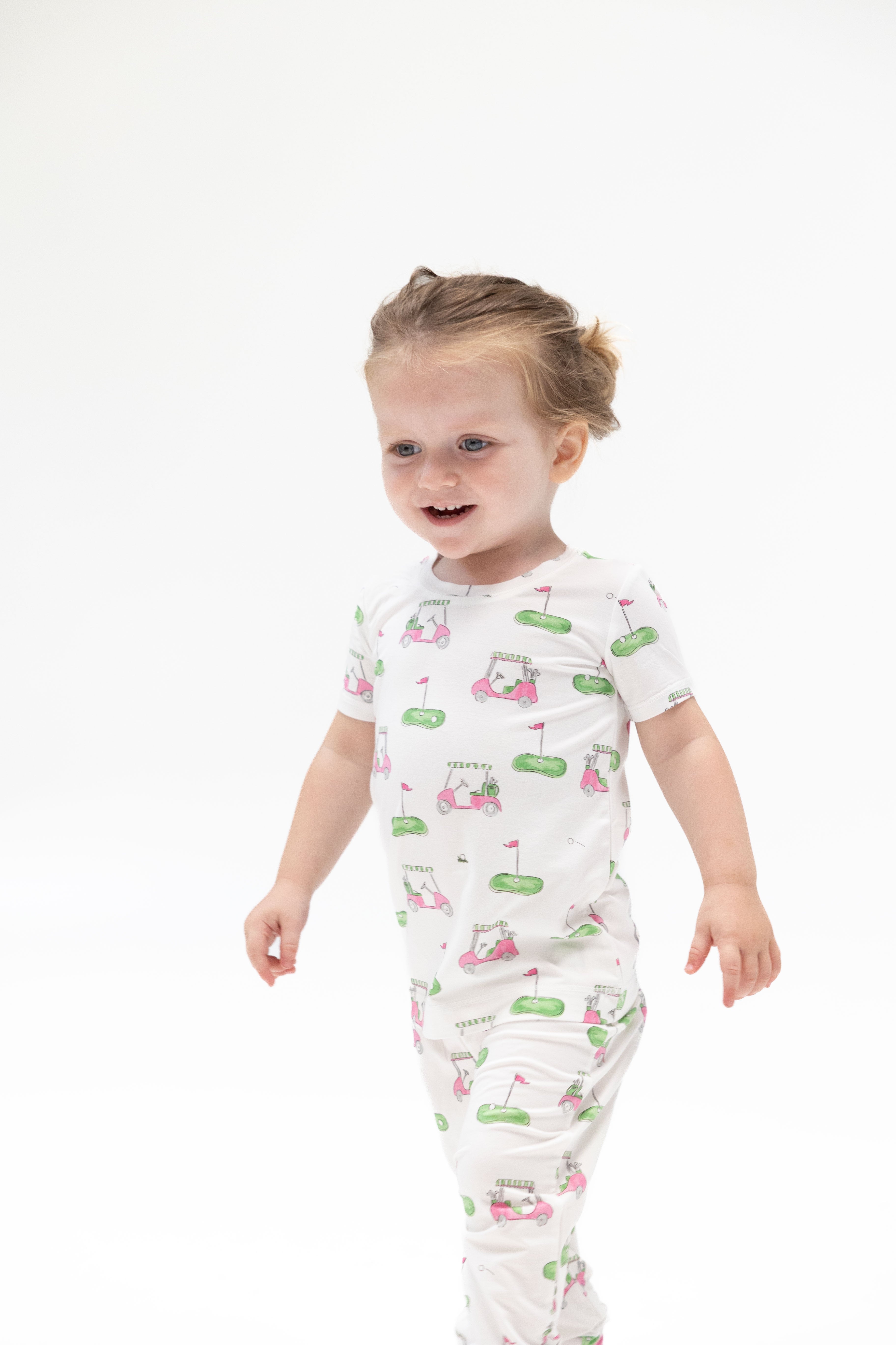 A toddler girl wearing a white short-sleeve footless pajama romper with a green and pink golf course and cart pattern, standing against a solid white background.