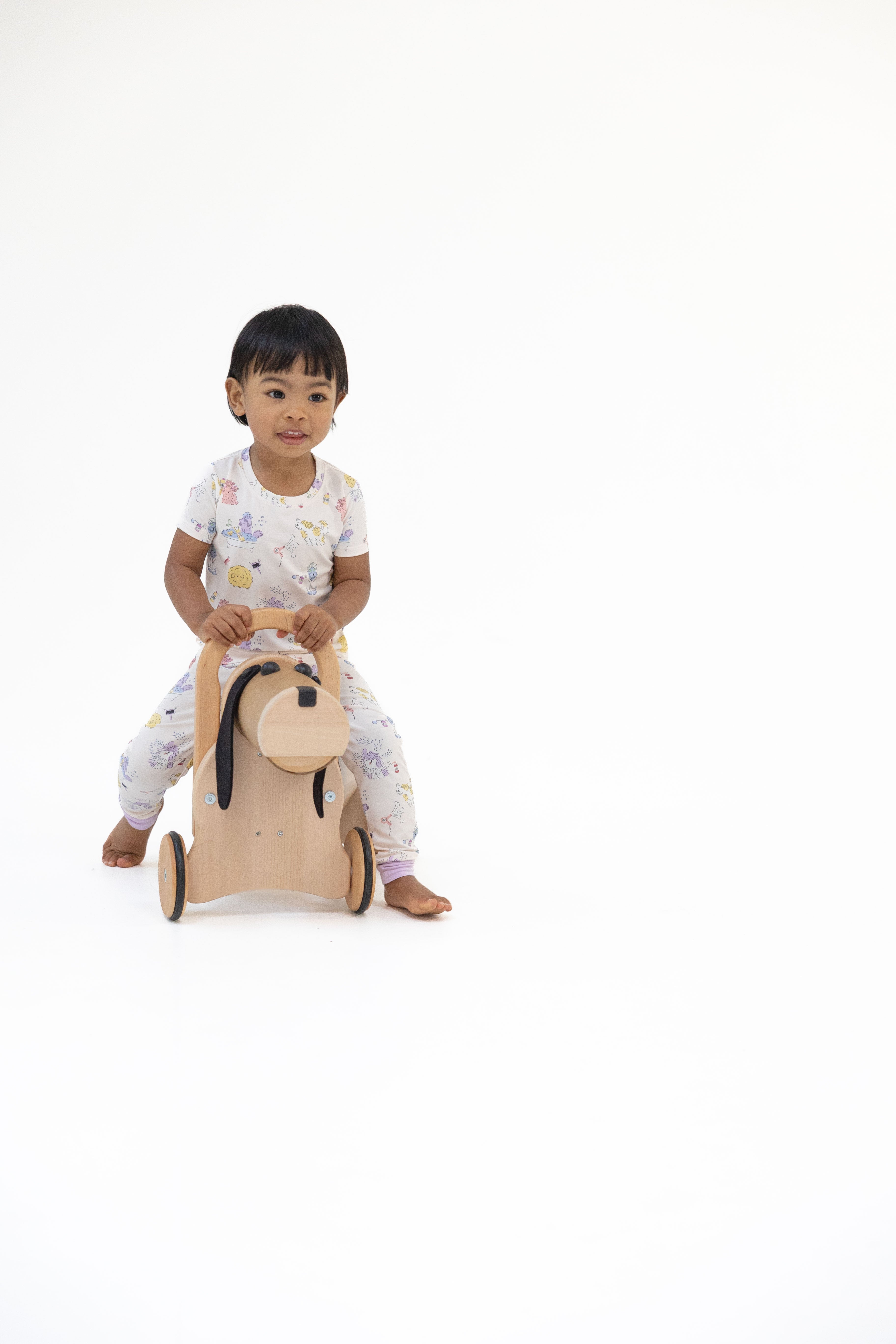 A toddler wearing a white doggy print pajama set sits on a wooden horse toy against a solid white background.