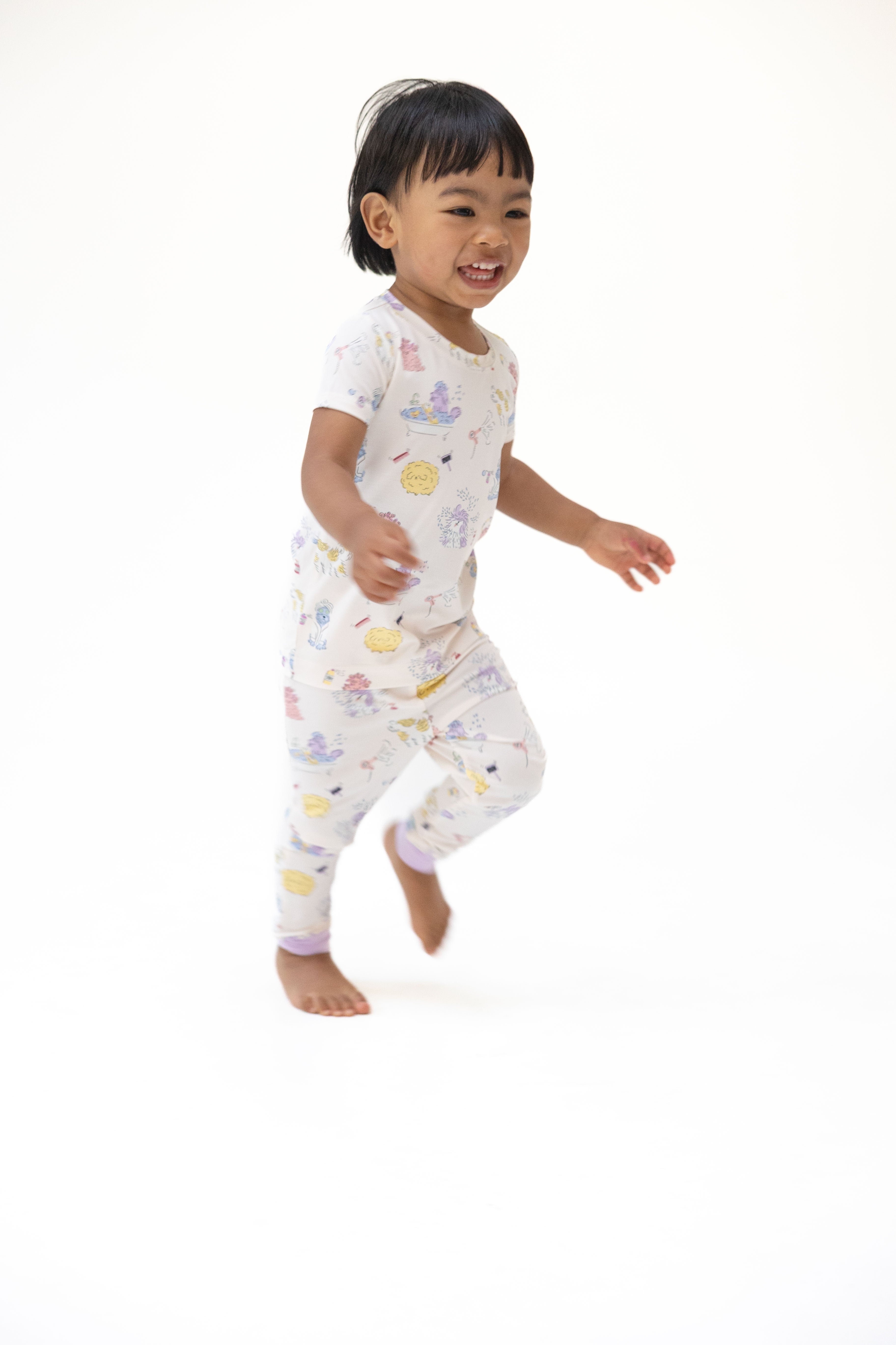 A smiling toddler running barefoot in a white short-sleeve pajama set with a pastel multicolor dog pattern, captured against a seamless white studio background.